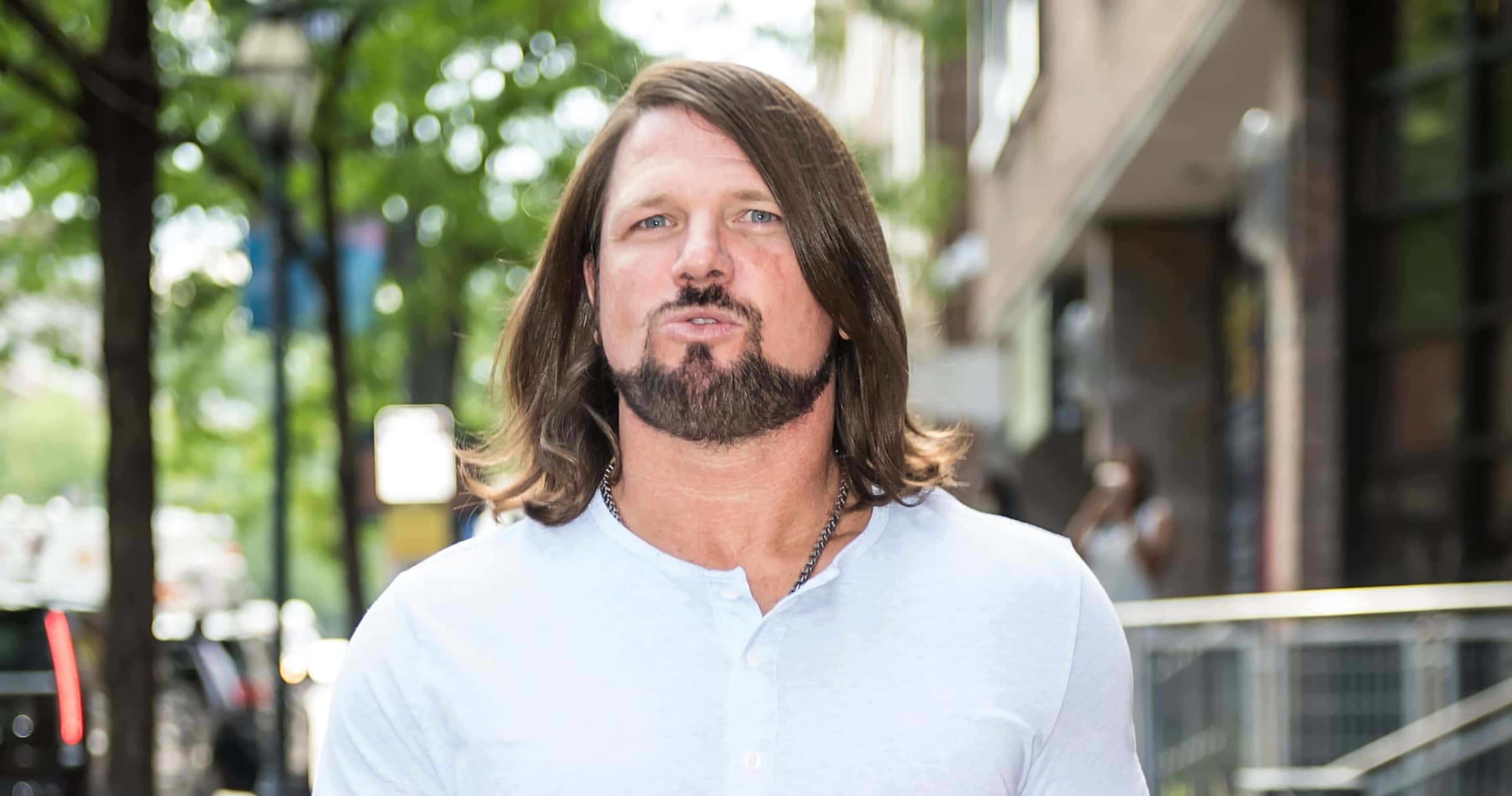AJ Styles 休閒裝扮 壁紙 — 一位留著長棕色頭髮和鬍鬚的男人自信地站在陽光普照的城市人行道上，周圍環繞著樹木和建築物。