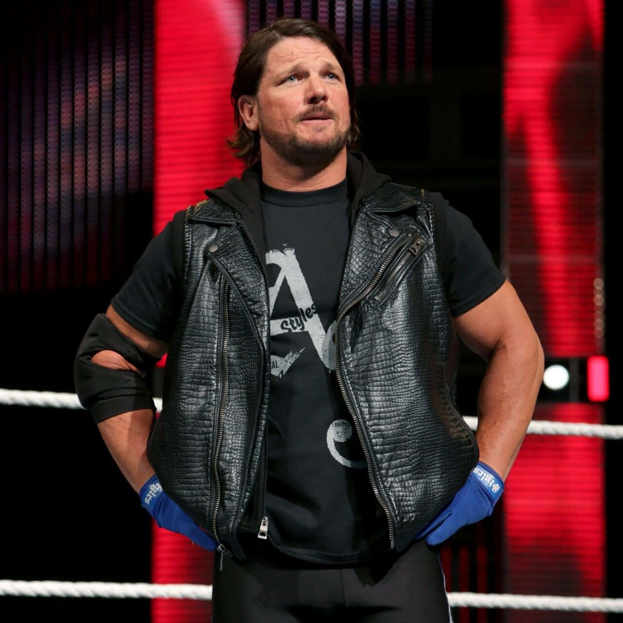 AJ Styles 八次世界冠軍 壁紙 — 摔角手AJ Styles自信地站在摔角擂台上，身穿黑色皮背心和藍色手套，背景為紅色燈光。