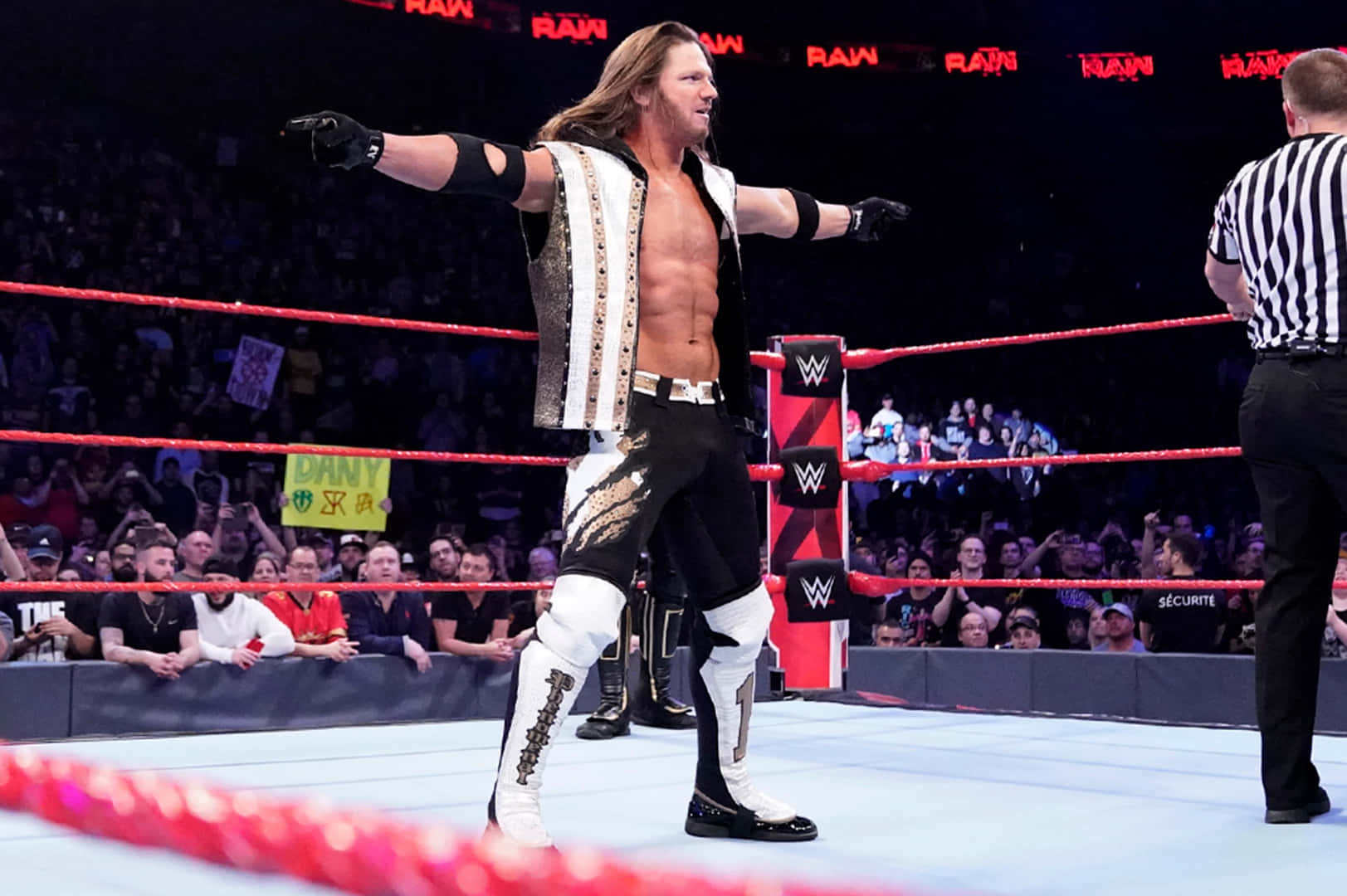 AJ Styles：摔角界的熱情與表現力化身 壁紙 — 一位肌肉發達的摔角手身穿黑白服裝，在摔角場上張開雙臂勝利地站立，裁判在旁觀看，背景中粉絲歡呼。