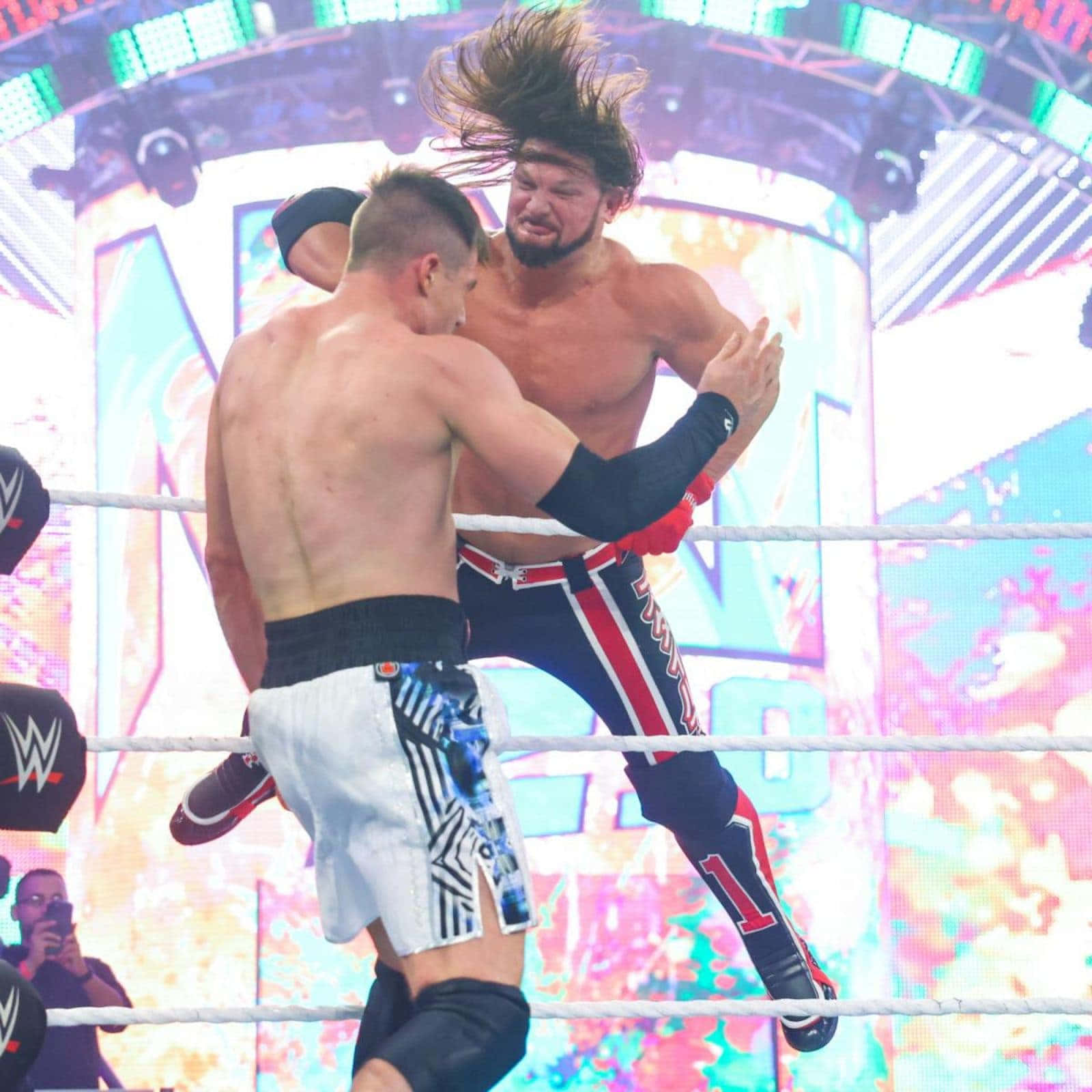 AJ Styles 職業摔角手 壁紙 — 兩名摔角手在擂台上碰撞，其中一人凌空施展招式，背景是充滿活力的數位畫面。