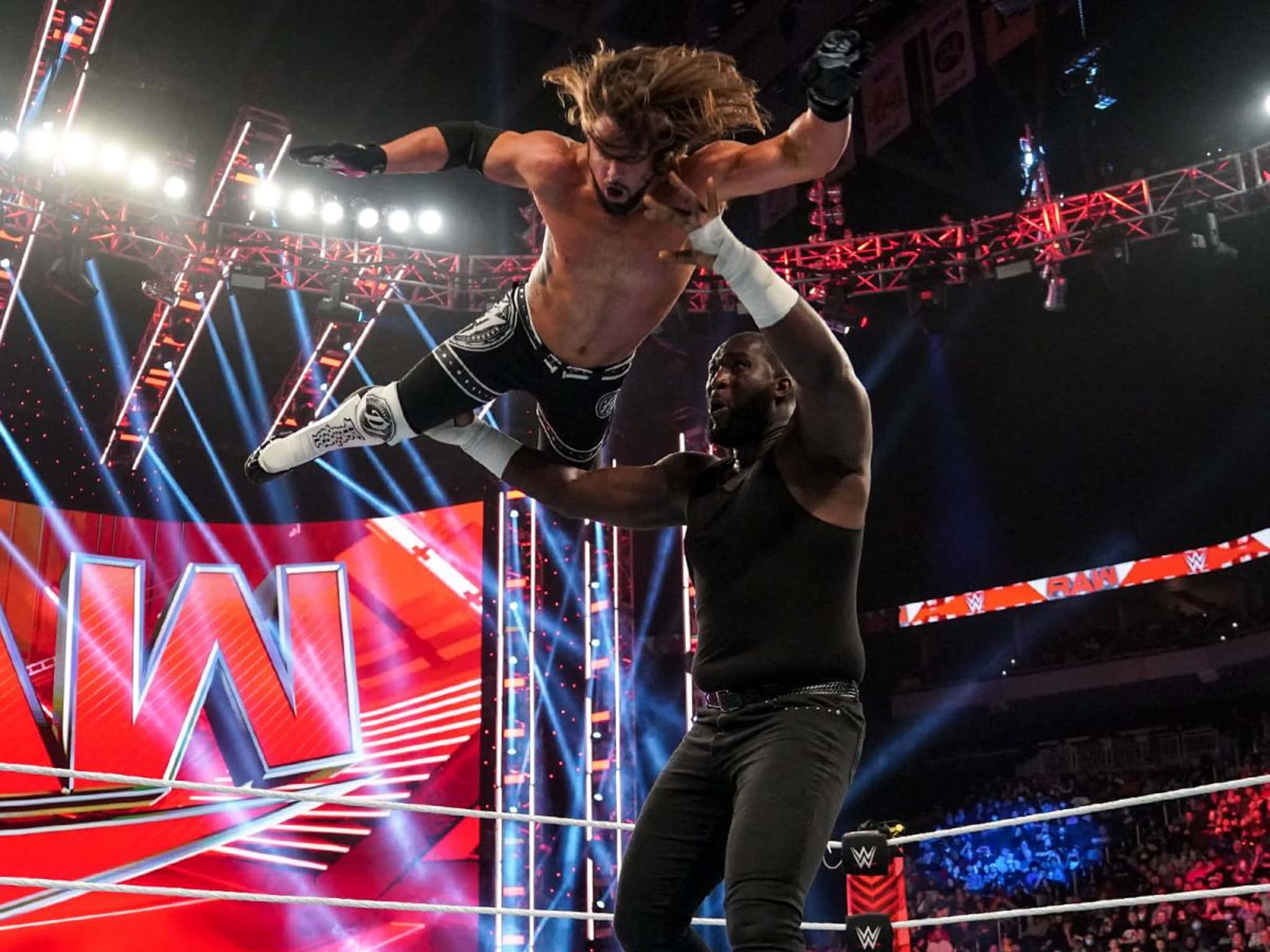 AJ Styles 與 Omos WWE Raw 壁紙 — 一名摔角手在明亮的舞台燈光下，於摔角擂台上將另一人高舉過頭頂。