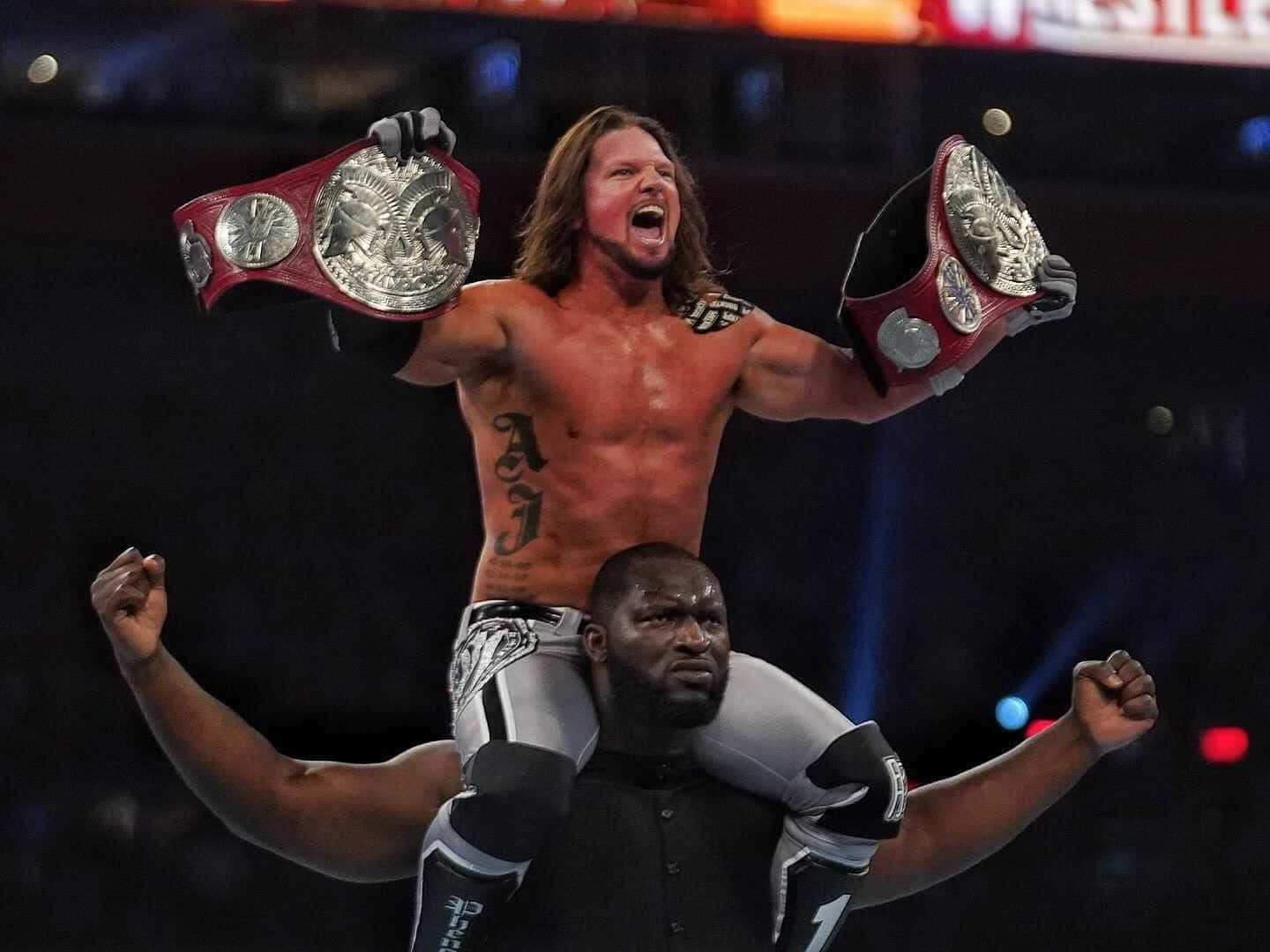 AJ Styles 與 Omos 庆祝勝利 壁紙 — 摔角手AJ Styles在燈光明亮的場館中，站在拍檔的肩膀上，高舉兩條冠軍腰帶，勝利姿態盡顯。
