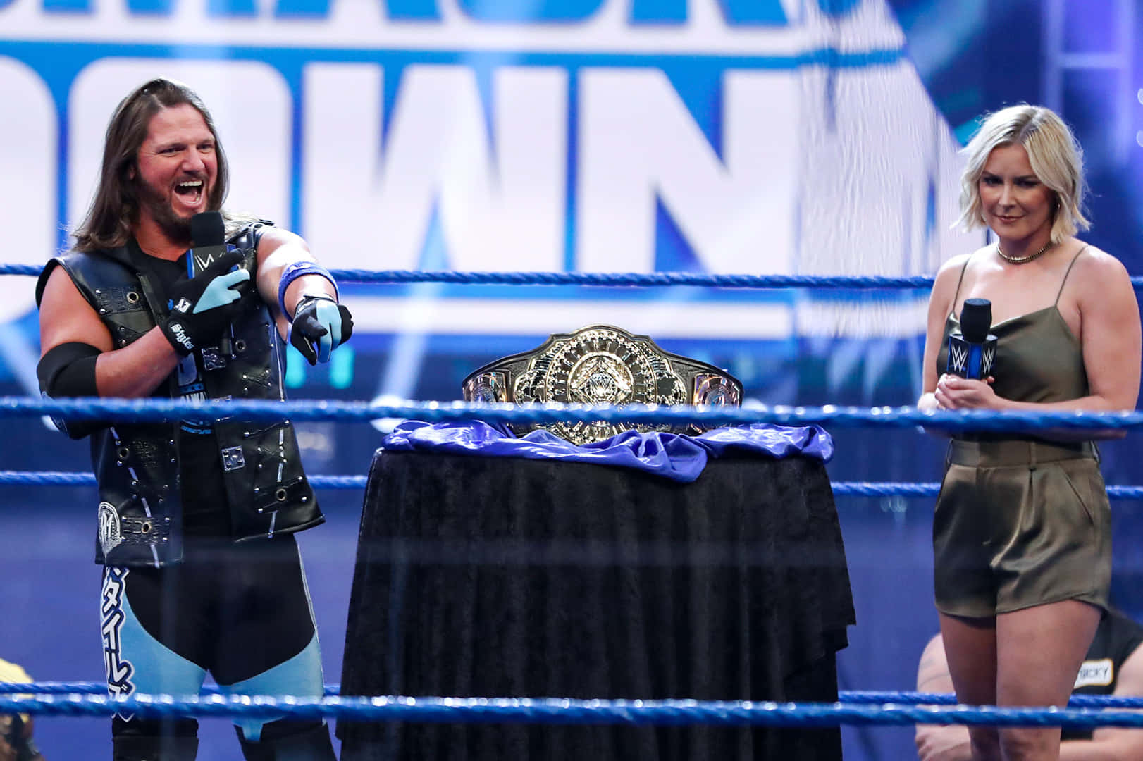 AJ Styles 與Renee Young 壁紙 — 身穿黑色和藍色裝備的摔角手在女主持人旁邊勝利地咆哮，冠軍腰帶放在擂台上的桌子上。