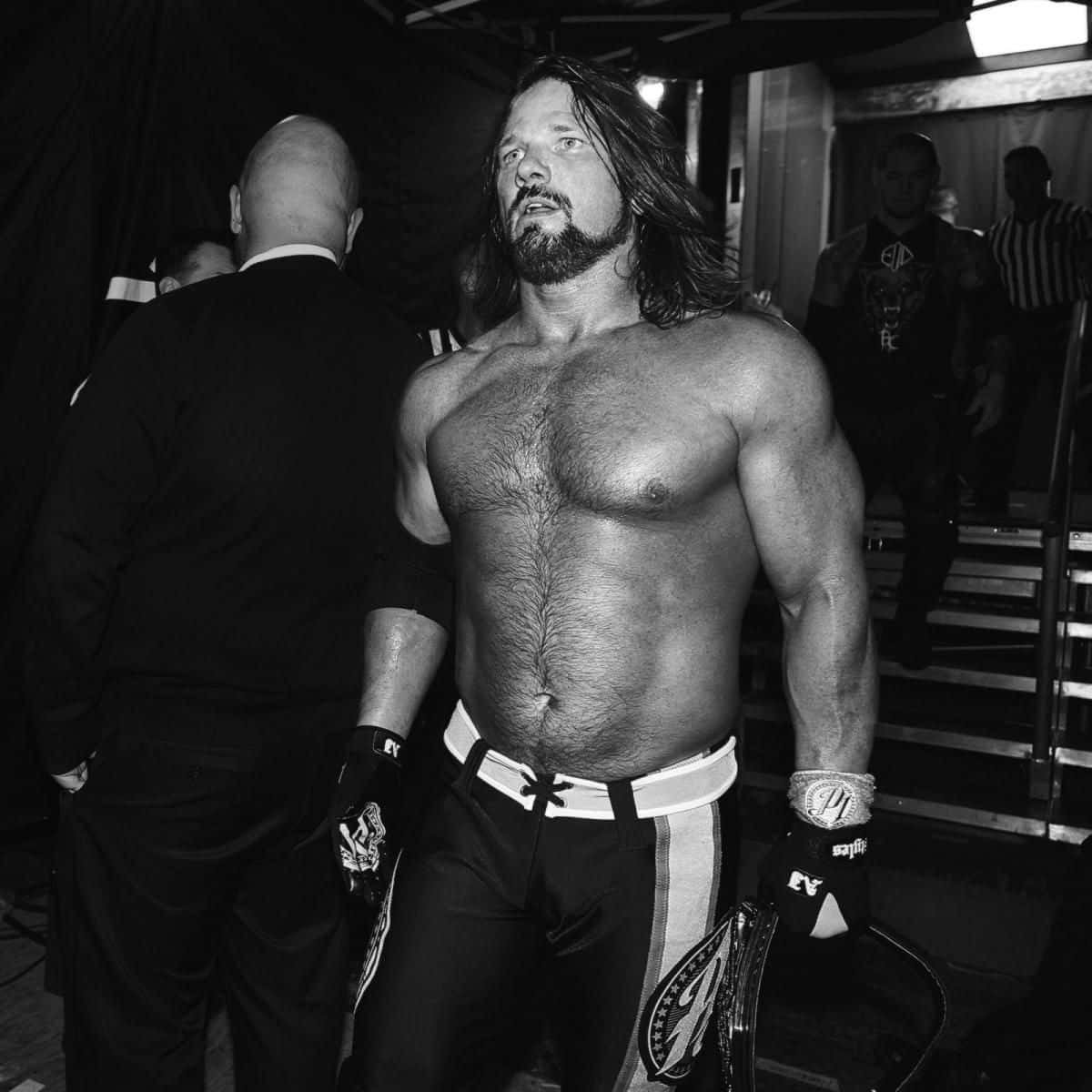 AJ Styles 黑白影像 壁紙 — 一位肌肉發達、長髮鬍鬚的摔角手，赤裸上身站在後台，在戲劇性的黑白燈光下手持冠軍腰帶。