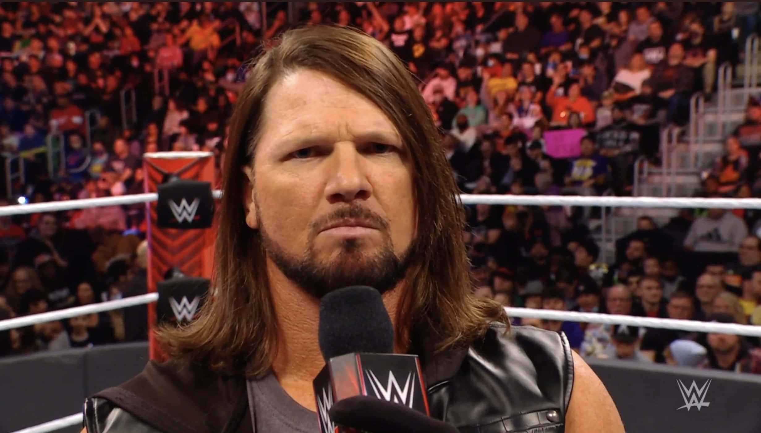 AJ Styles特寫鏡頭 壁紙 — WWE摔角手留著長棕色頭髮和鬍鬚，在摔角場內對著麥克風說話，身後是擠滿的人群。