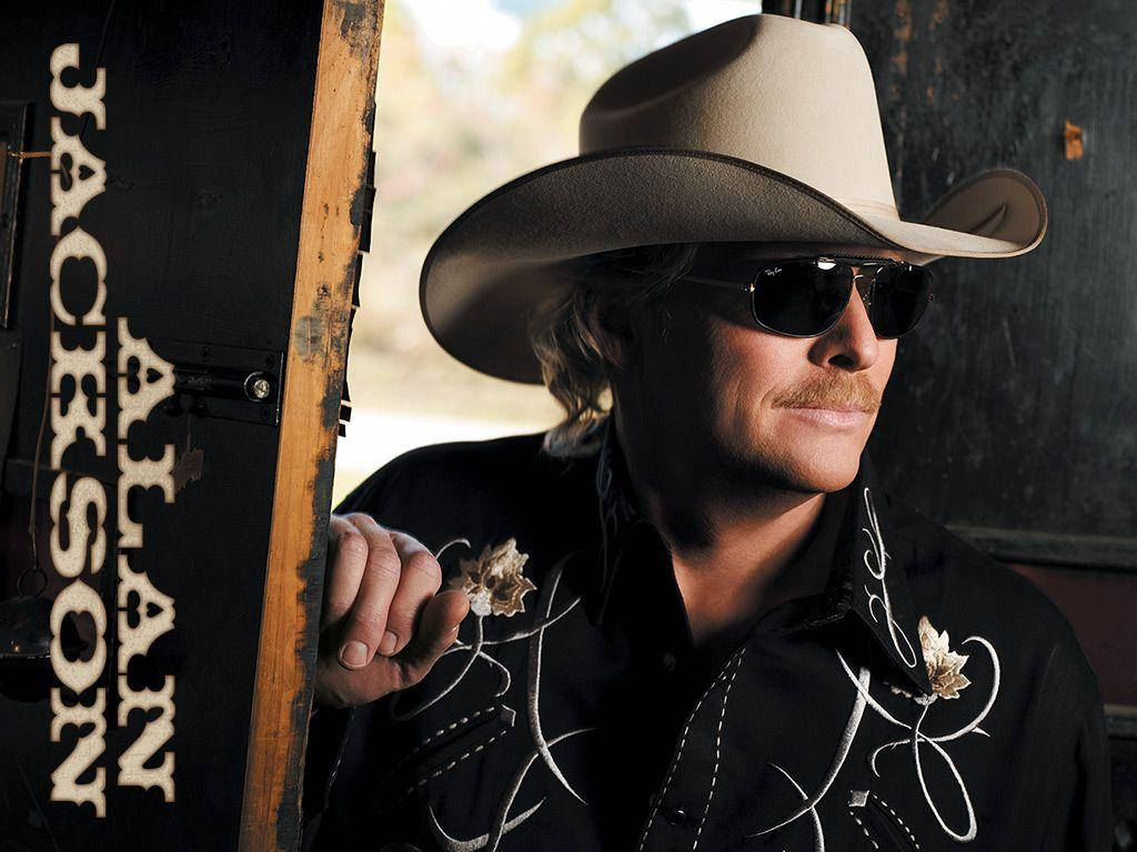 Alan Jackson 桌布 壁紙 — 一個戴著牛仔帽和太陽眼鏡的男人，靠著一塊寫有 'ALAN JACKSON' 的風化標誌，眼神自信。