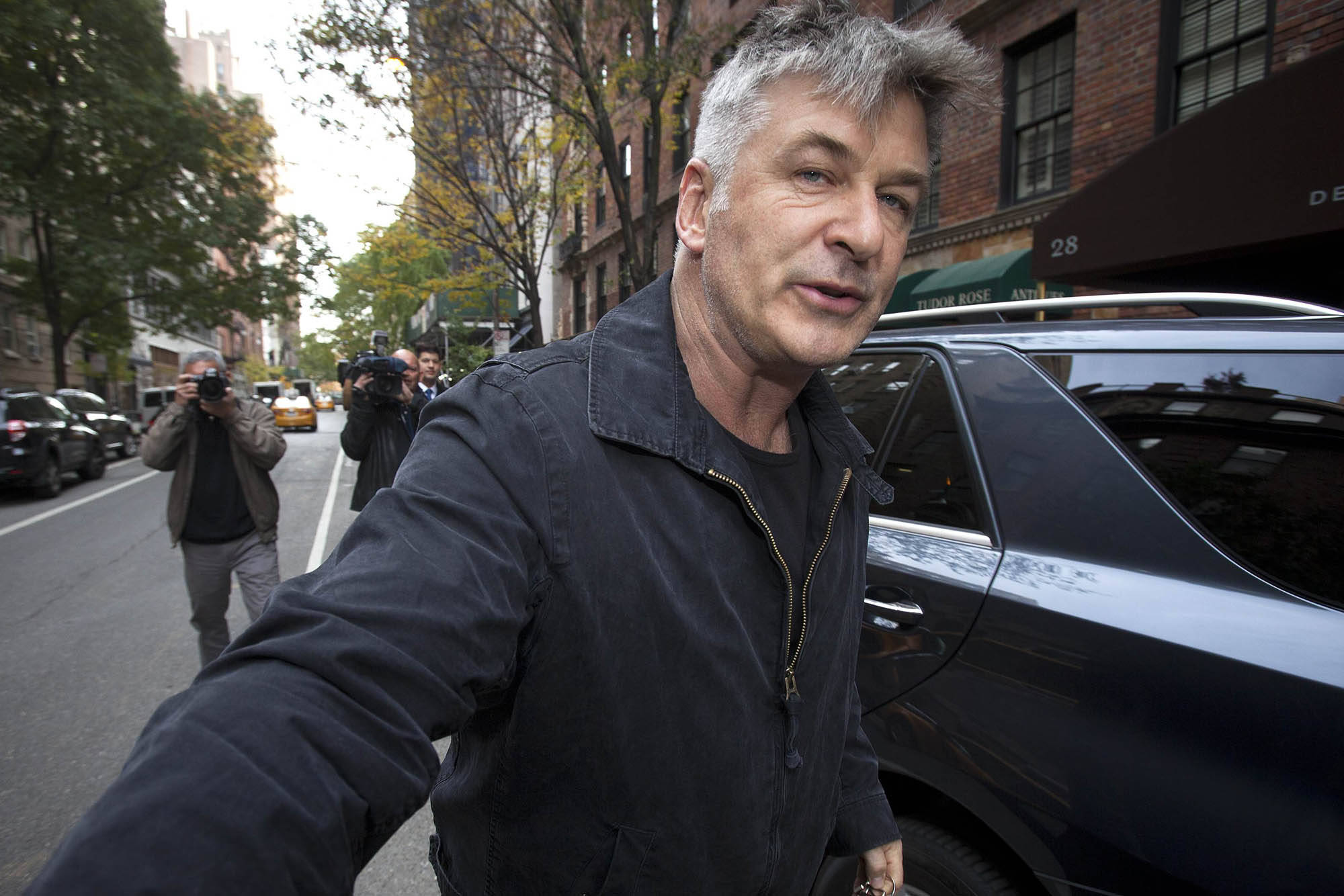 Alec Baldwin 被狗仔拍攝 壁紙 — 一位灰髮男子在晴朗的日子裡沿著城市街道行走，經過攝影師和停泊的汽車。