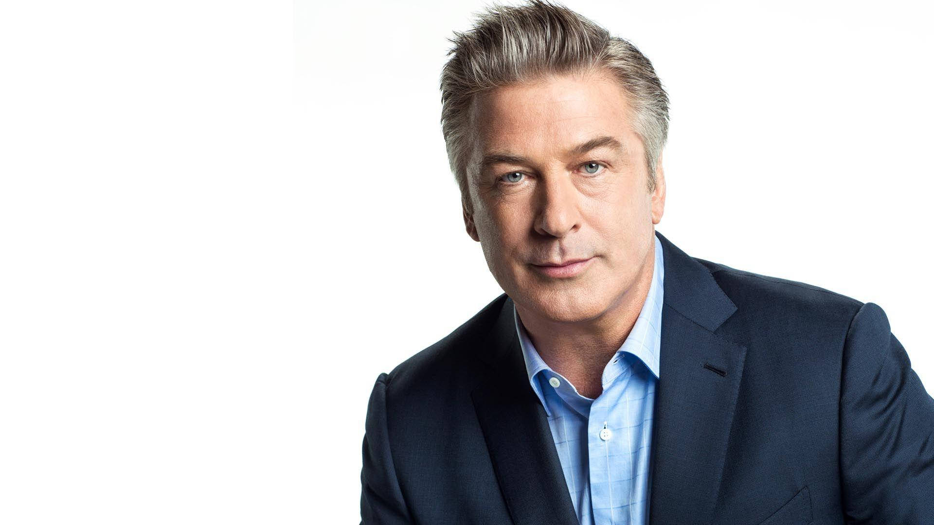 Alec Baldwin 訪問拍攝 壁紙 — 一位自信嘅中年男士，灰白頭髮，身穿深色西裝同淺藍色恤衫，背景係純白色。