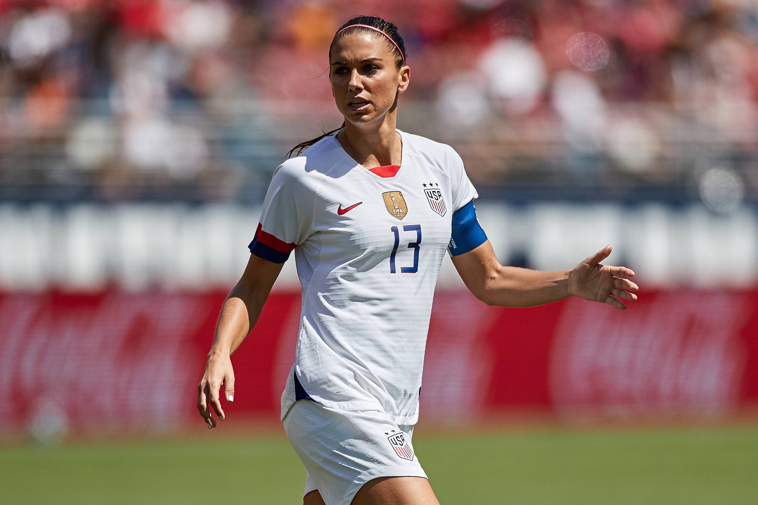 美國隊 Alex Morgan 壁紙 — 一位專注的女足球員身穿白色美國國家隊13號球衣，在充滿活力的綠色球場上進行日間比賽時做出手勢。
