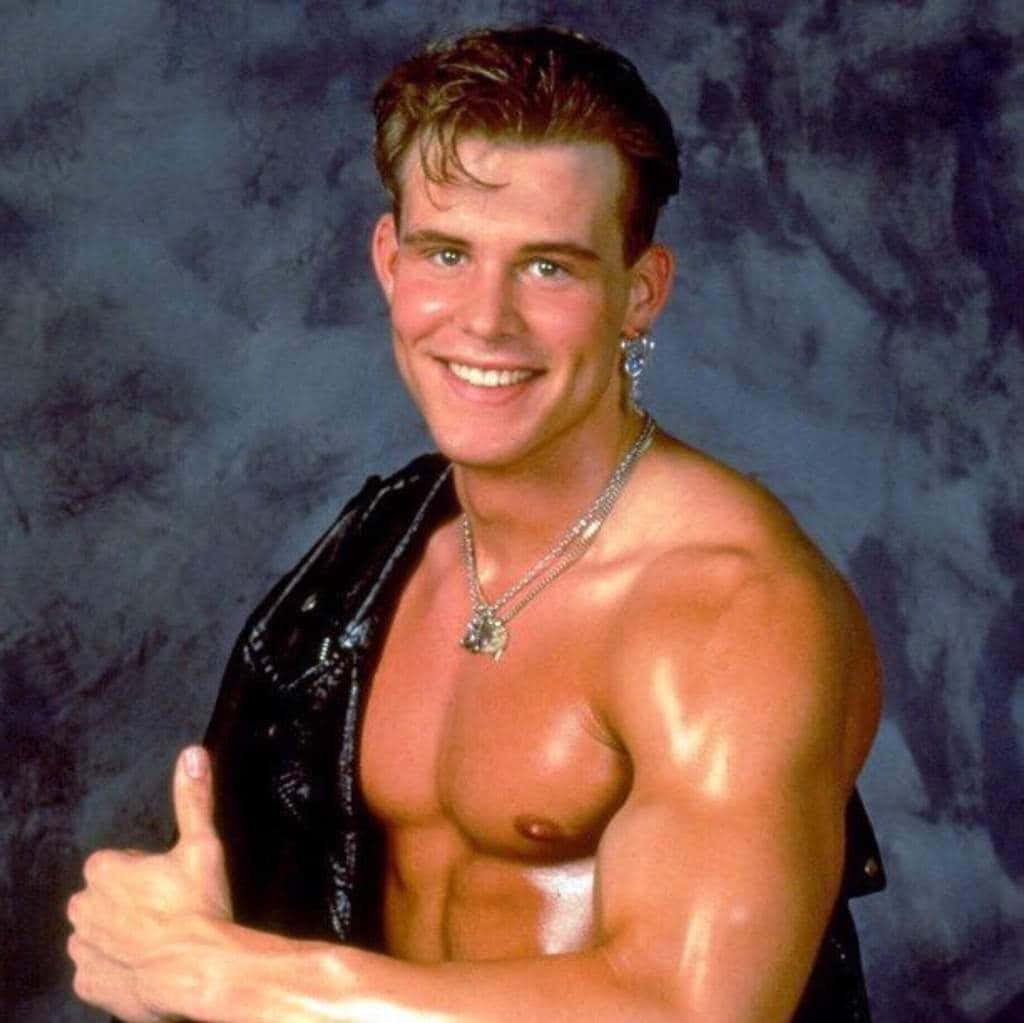 Alex Wright WCW 摔角才華 壁紙 — 一個肌肉發達、漂染頭髮、佩戴銀飾的男人，在帶有紋理的藍色背景下自信地豎起大拇指。