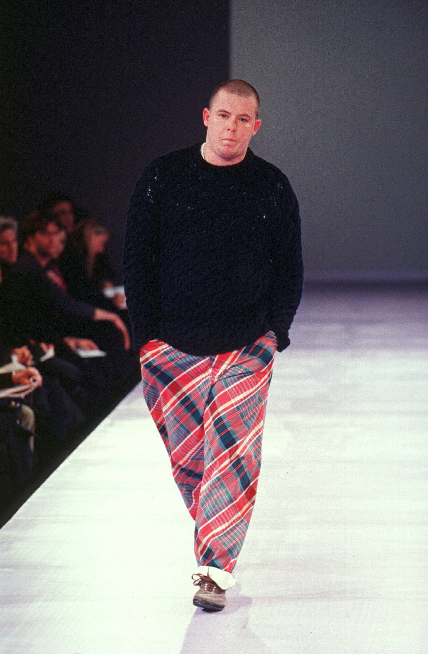 Alexander McQueen 身著 Comme Des Garçons 壁紙 — 模特兒在走秀，身穿黑色紋理毛衣和鮮明的紅藍格子褲，在明亮的攝影棚燈光下。