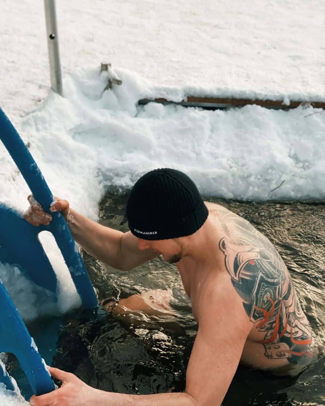 有紋身的男子在白雪皚皚的冬日景色中用藍色梯子從冰水中爬出。