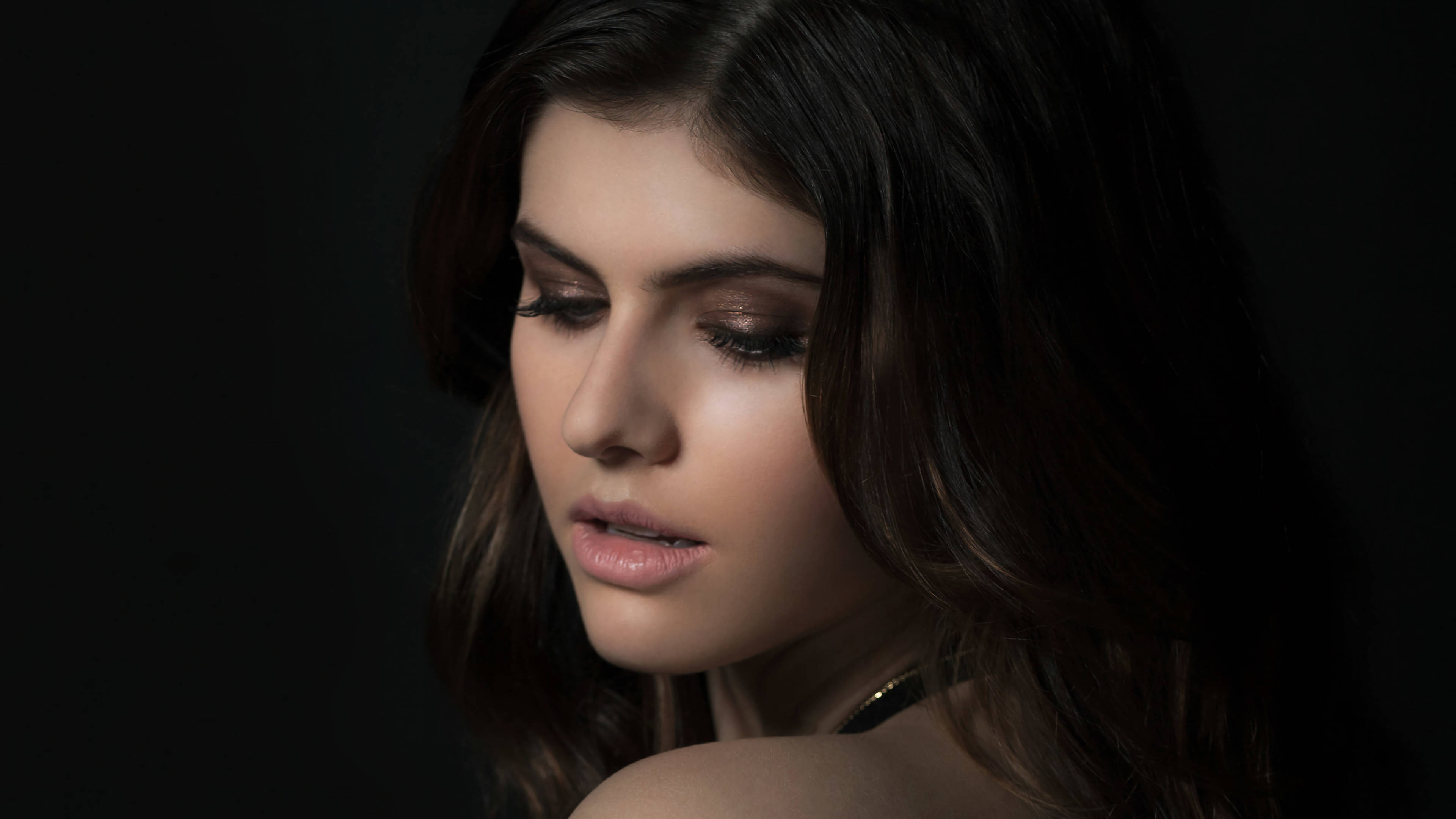女演員Alexandra Daddario 4K畫質 壁紙 — 一位深色頭髮、妝容柔和的女性在黑色背景和戲劇性燈光下向下凝視。