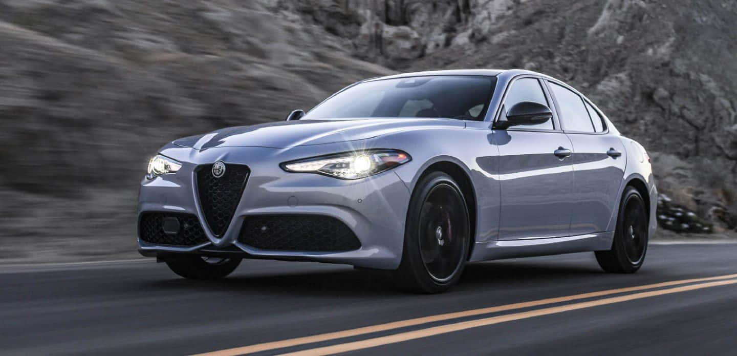 尊貴的Alfa Romeo 壁紙 — 一輛時尚的銀色阿爾法羅密歐轎車沿著蜿蜒的山路疾馳，背景是模糊的岩石懸崖。