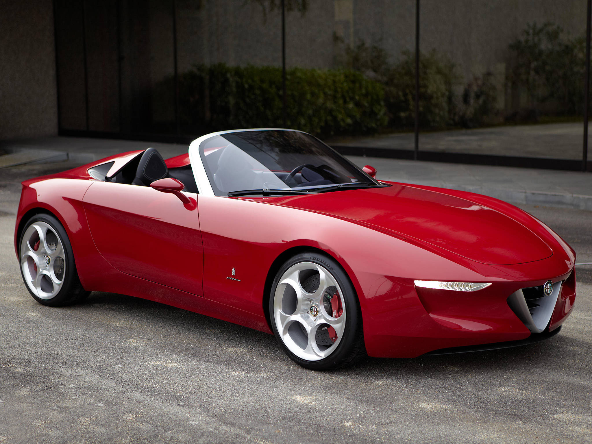 Alfa Romeo iPhone 停泊 壁紙 — 一輛亮紅色的愛快羅密歐Spider開篷車停在瀝青路上，背景有現代玻璃建築和綠植。