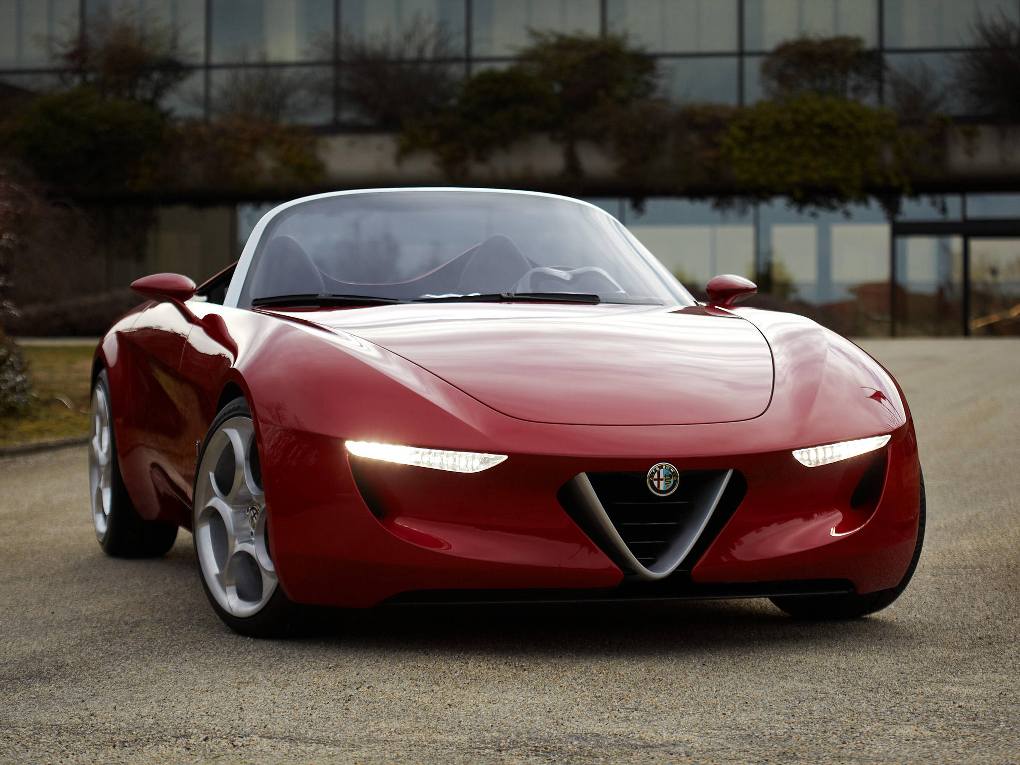 Alfa Romeo iPhone 頭燈 壁紙 — 一輛時尚的紅色愛快羅密歐概念車，帶有發光車頭燈，停在現代玻璃建築前的礫石上。