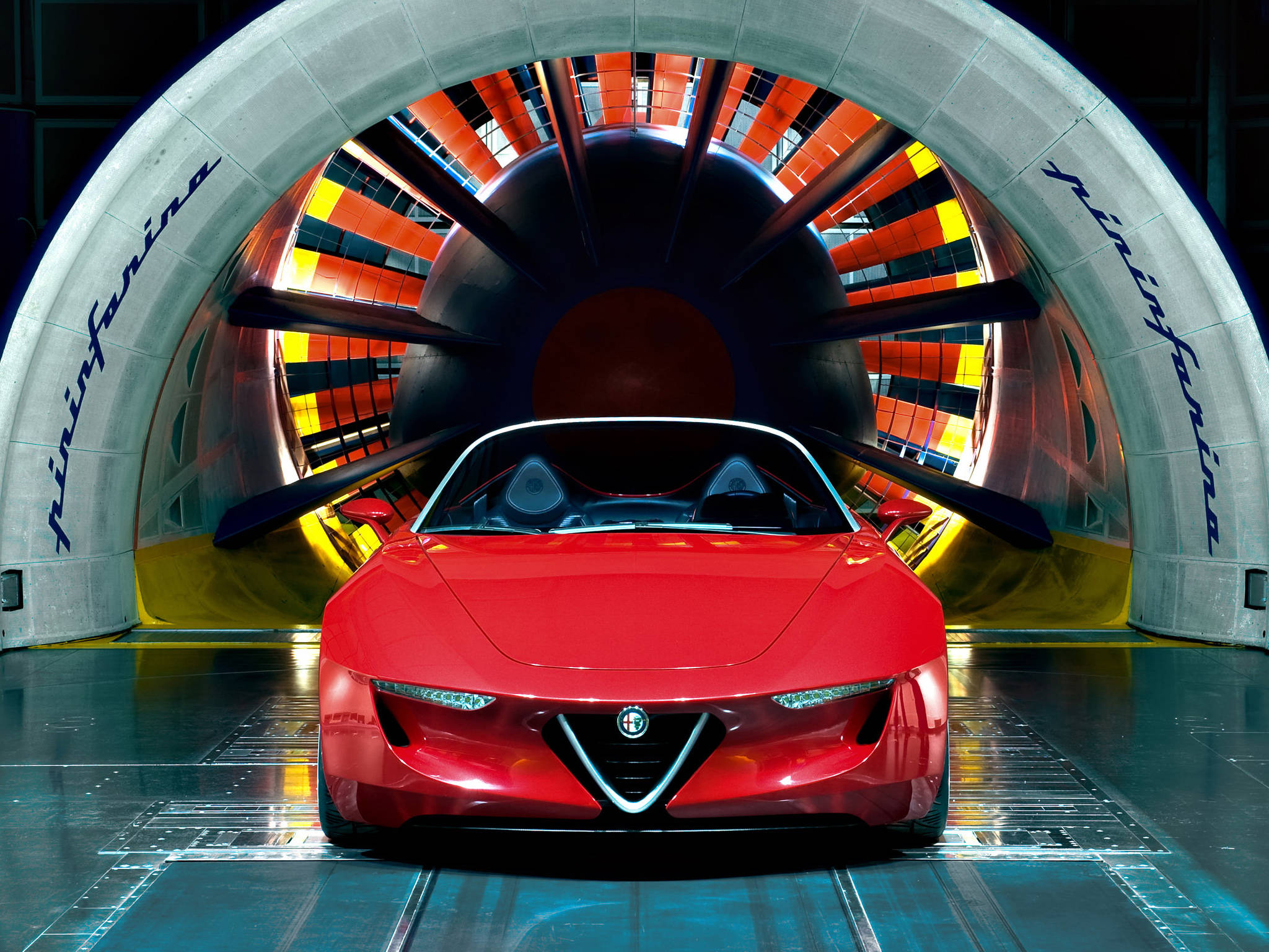 Alfa Romeo iPhone 紅色 壁紙 — 一輛時尚的紅色跑車位於高科技風洞中央，背後有鮮豔的橙色和黃色葉片旋轉。