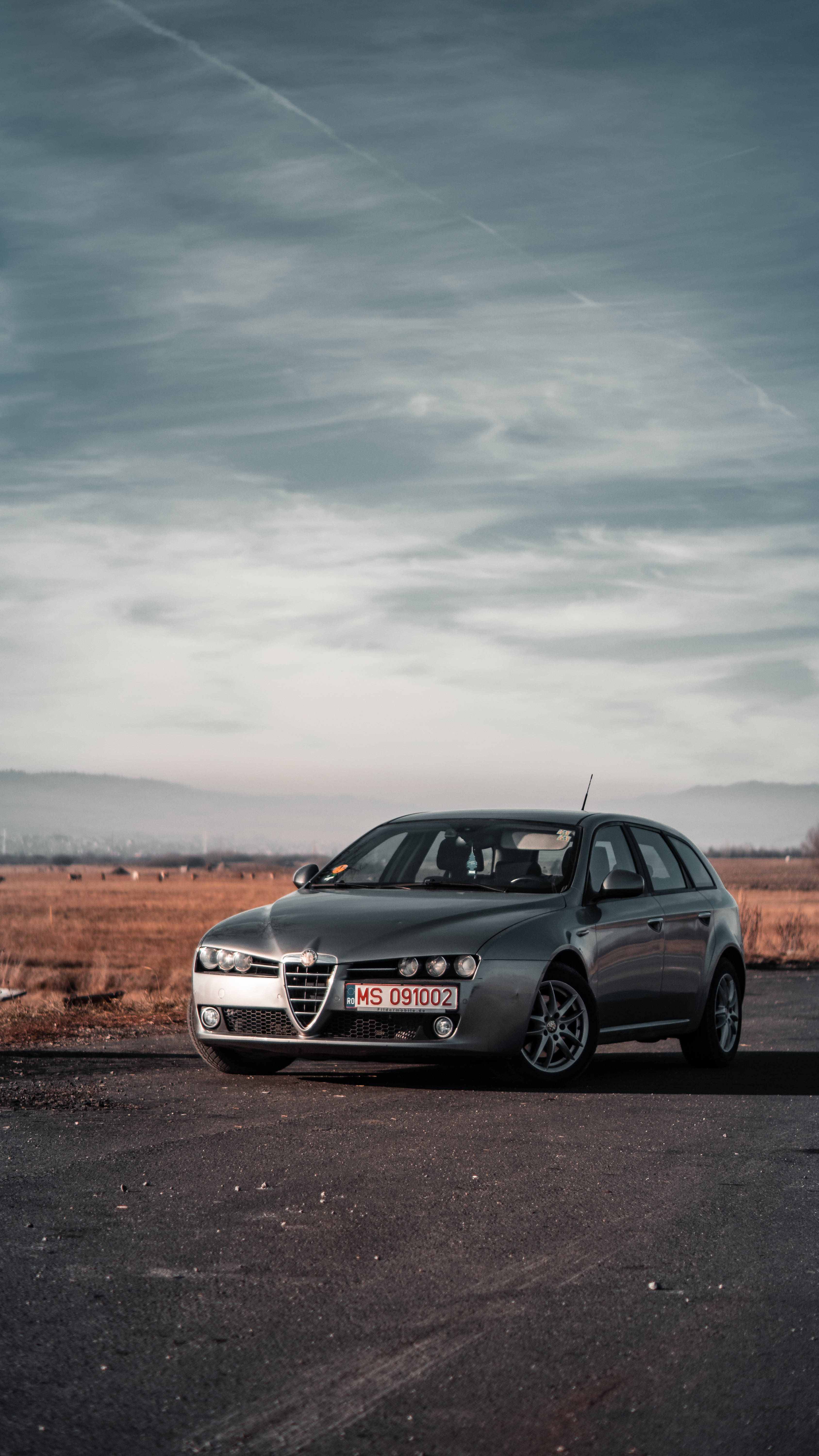 Alfa Romeo iPhone 灰色 壁紙 — 一架時尚的銀色愛快羅密歐旅行車停泊在瀝青路上，背景是廣闊多雲的天空和遠山。
