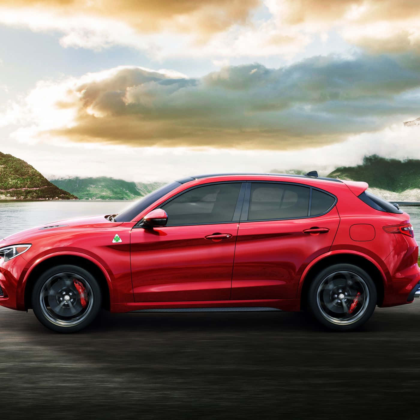 「Alfa Romeo Stelvio 的迷人之美」 壁紙 — 一輛鮮紅色的Alfa Romeo Stelvio SUV沿著湖邊道路行駛，背景是霧氣繚繞的山脈和金色的雲彩。