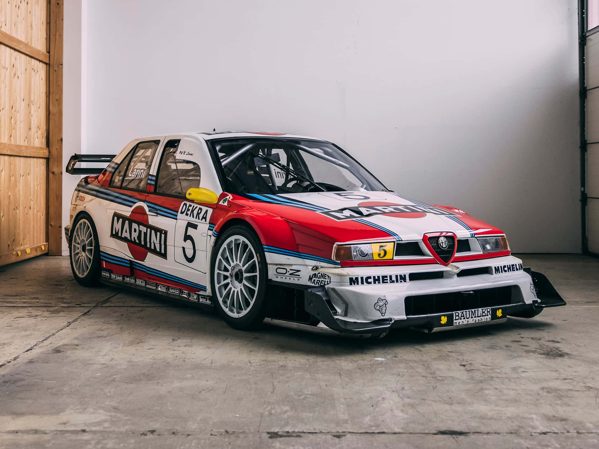 Alfa Romeo 時光不變的優雅設計 壁紙 — 一輛帶有馬天尼塗裝的復古愛快羅密歐賽車停在混凝土車庫中，展示大膽的紅白藍條紋和贊助商貼紙。