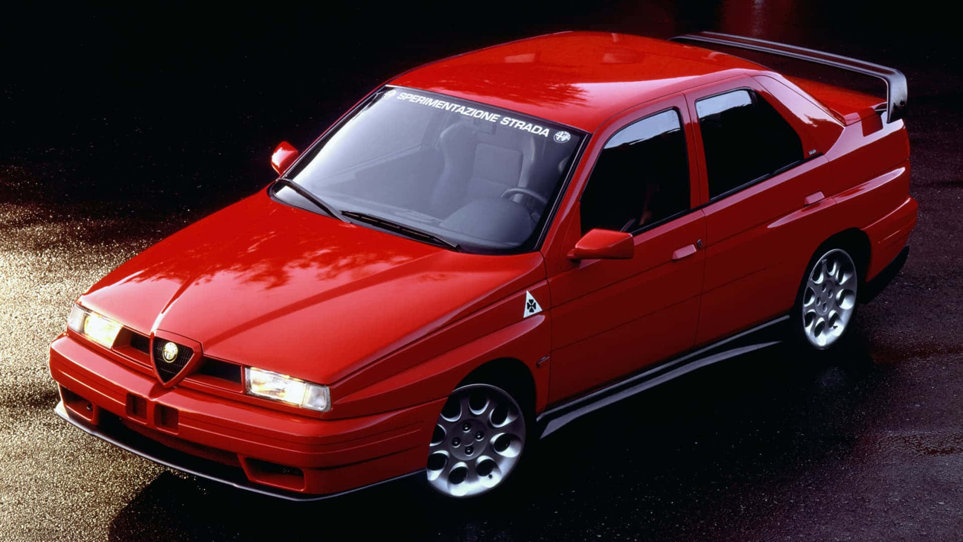 Alfa Romeo，經典設計，永恆傳承 壁紙 — 一輛光澤的紅色Alfa Romeo 155 V6 Sport轎車在潮濕的瀝青路面上於攝影棚燈光下閃耀。