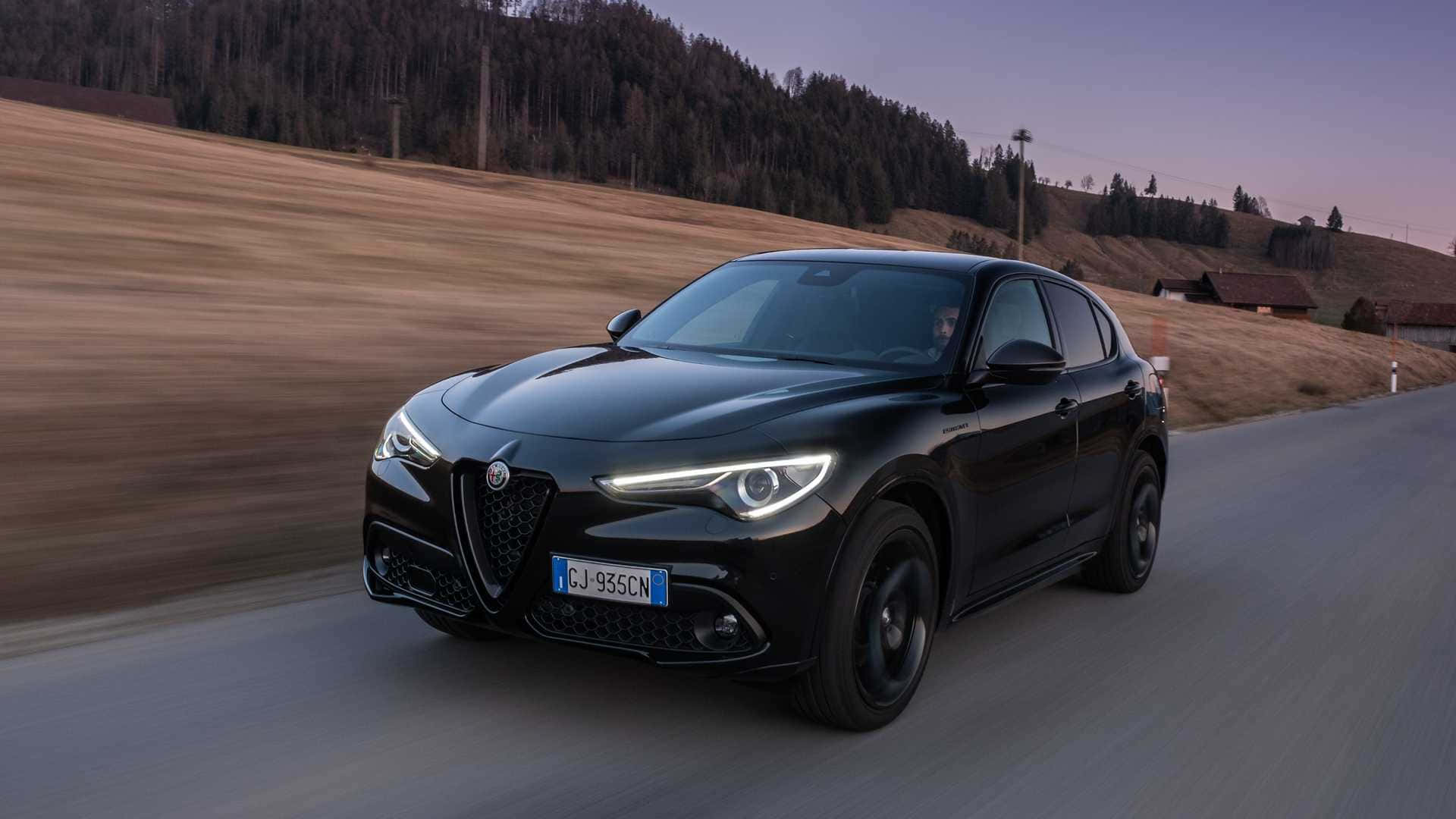 Alfa Romeo：經典跑車自信 壁紙 — 一輛時尚的黑色Alfa Romeo SUV在黃昏時分沿著蜿蜒的鄉村道路疾馳，背景是模糊的金色田野和森林覆蓋的山丘。