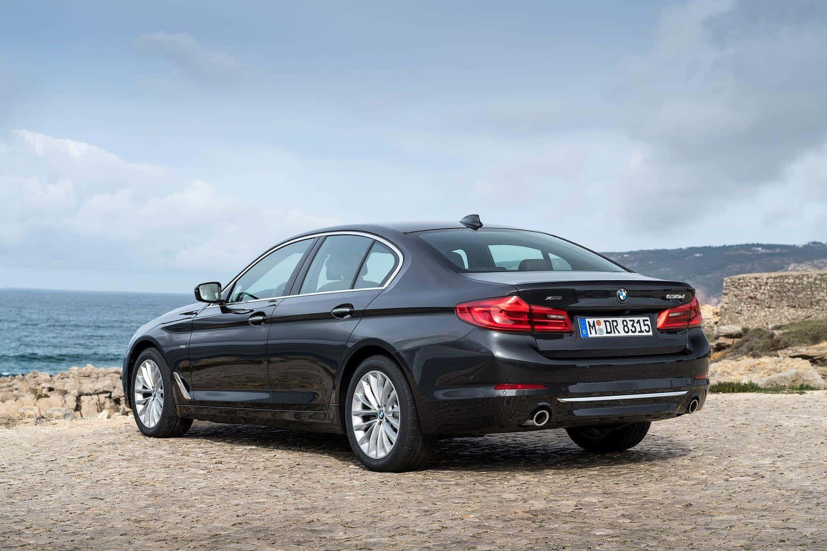 Alfa Romeo車輛停駐於義大利古堡前 壁紙 — 一架時尚的黑色BMW 5系轎車停在鵝卵石懸崖上，俯瞰著多雲天空下的海洋。