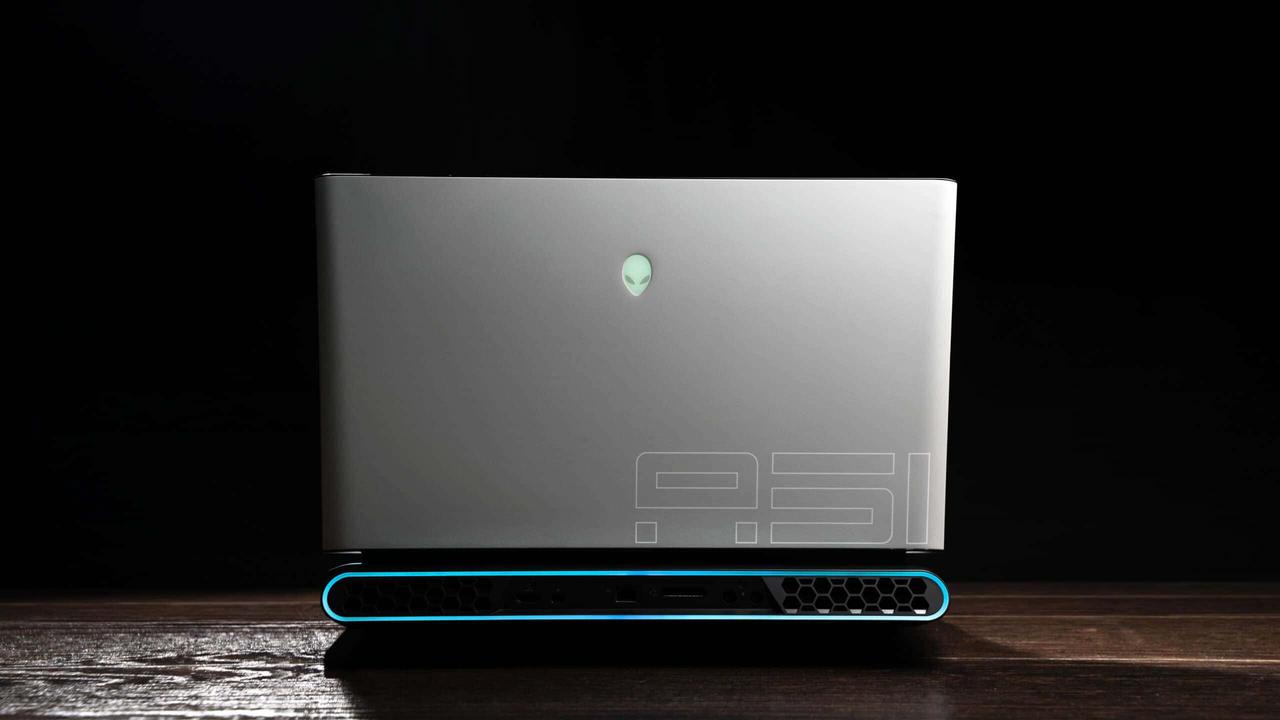 以 Alienware 挖掘您的遊戲潛能 壁紙 — 一部時尚的銀色Alienware筆記型電腦，帶有發光的藍色點綴，關閉在黑色背景下的深色木質表面上。