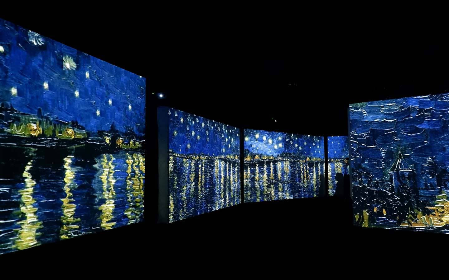 梵高 Alive 博物館 壁紙 — 一個黑暗的畫廊展示了梵高《星夜》的沉浸式數碼投影，鮮豔的藍色和發光的黃色在水面上反射。