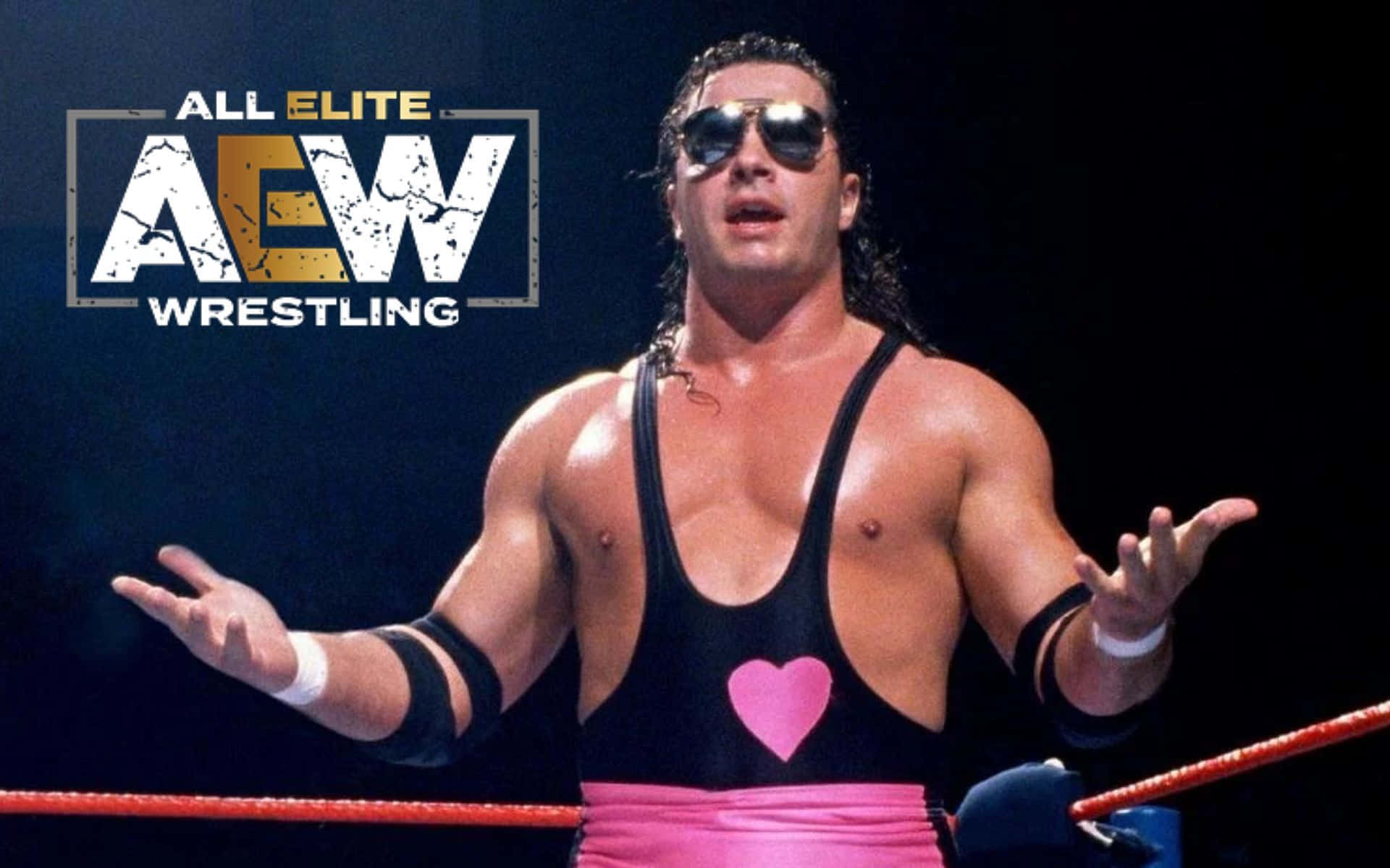 傳奇奧文·哈特於All Elite Wrestling擂台 壁紙 — 一位肌肉發達的摔角手穿著黑色背心，上面有粉紅色心形圖案和粉紅色緊身褲，戴著太陽眼鏡站在摔角場上，在明亮的燈光下戲劇性地做手勢。