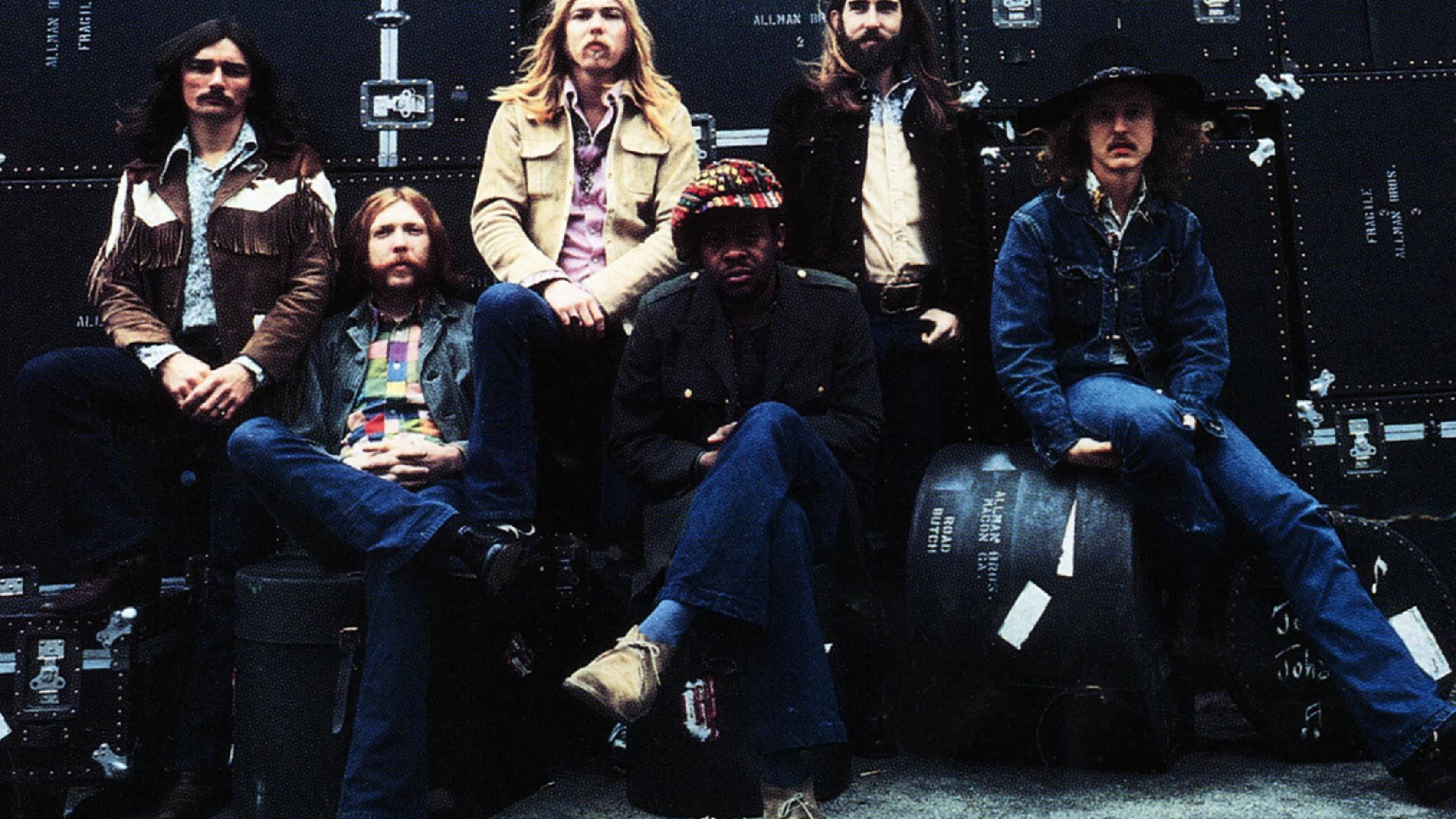 Allman Brothers Band 原始成員 壁紙 — 六位身穿1970年代搖滾服飾的音樂人，在黑暗的巡迴巴士背景下，隨意地坐在設備箱上擺姿勢。
