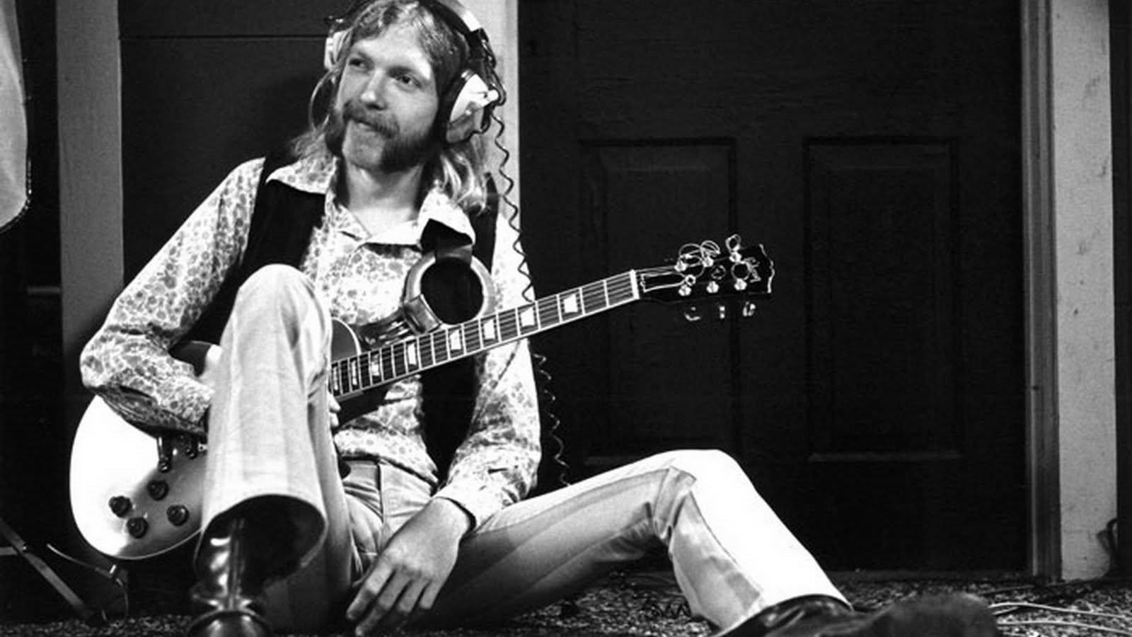 Allman Brothers Band 主音吉他手 Duane Allman 壁紙 — 一位留鬍鬚的男人戴著耳機盤腿而坐，手裡拿著結他，身後是昏暗的房間和木門。