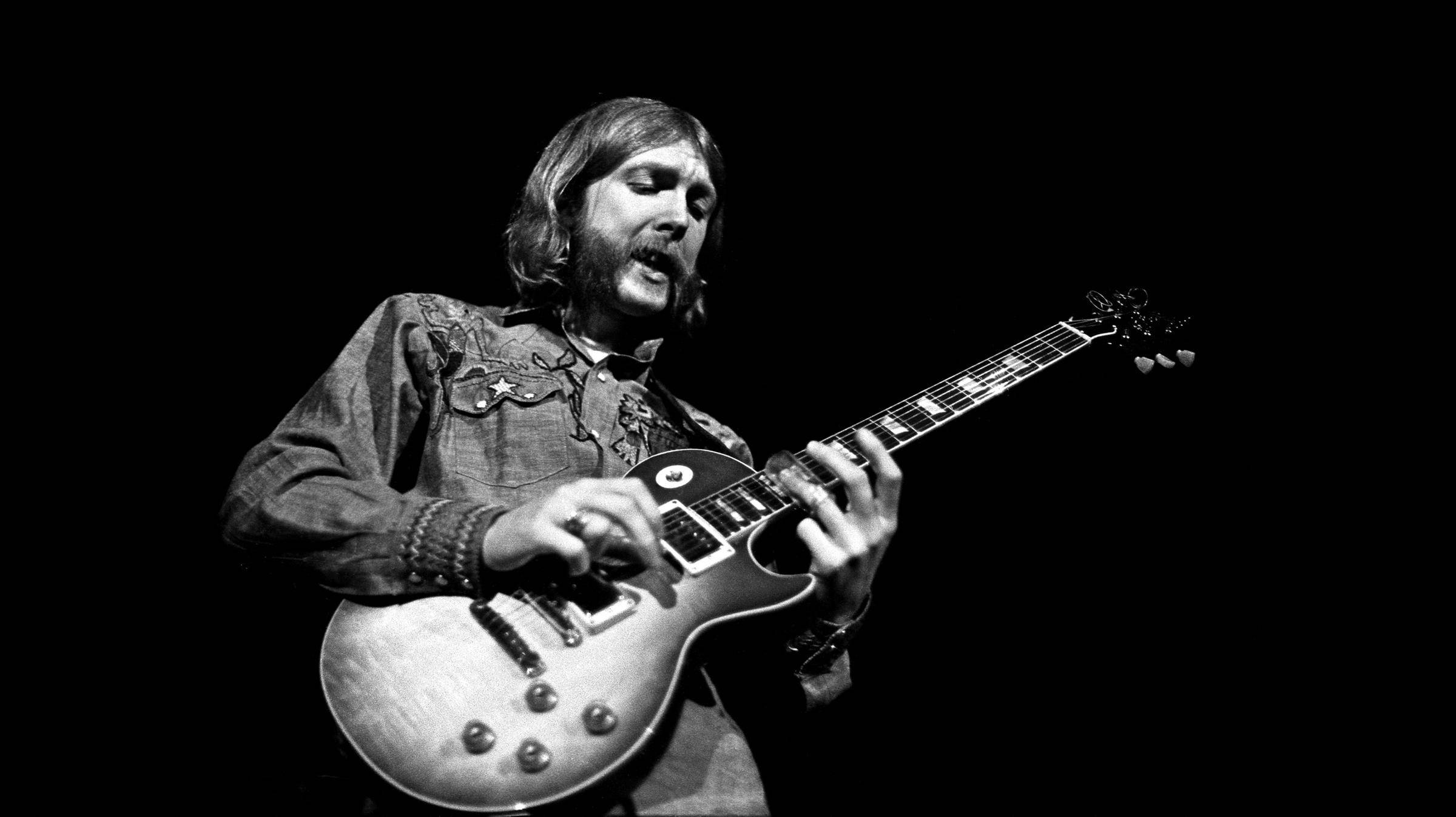 Allman Brothers Band 的 Gregg Allman 弹吉他 壁紙 — 一張黑白照片，一位鬍鬚音樂家在舞台上熱情地彈奏Gibson Les Paul電結他。