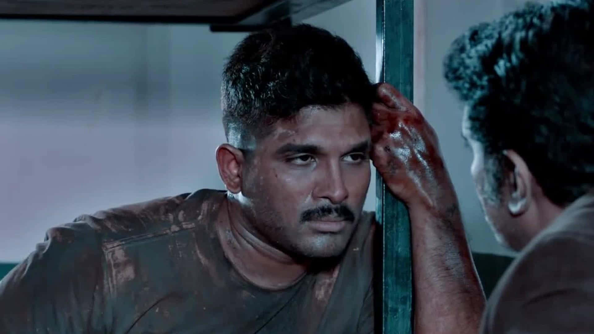 Allu Arjun在《Surya The Soldier》中的精彩演出 壁紙 — 一個手上有血的男人按壓金屬棒，同時在昏暗的房間裡凝視著另一個男人。
