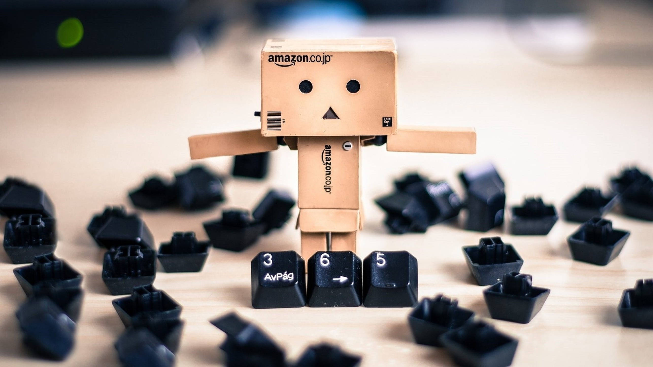 Amazon Boxman 與鍵盤鍵帽 壁紙 — 一個紙板亞馬遜機械人站在木桌上散落的黑色鍵盤按鍵之間。