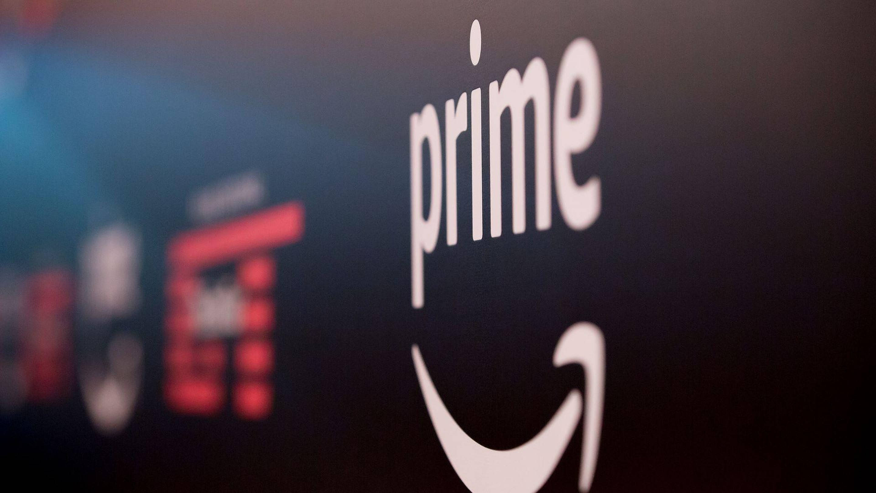 Amazon Prime標誌 壁紙 — 白色Amazon Prime標誌特寫，帶有微笑箭頭，背景為深色，遠處有模糊的紅色和藍色燈光。