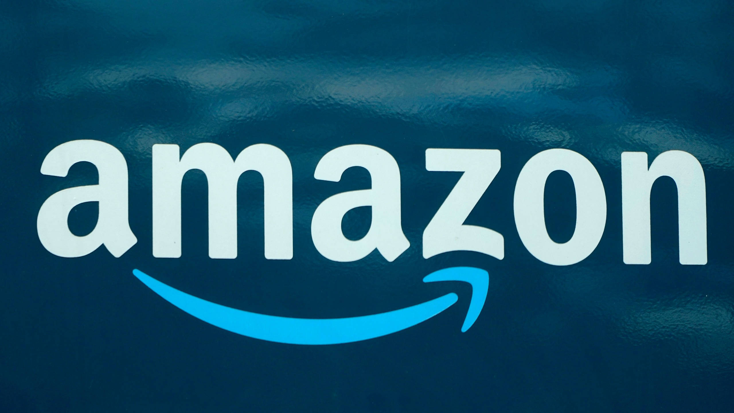 Amazon 水藍色背景 壁紙 — 亞馬遜標誌，白色和藍色文字，帶微笑箭頭，置於有紋理的深藍色背景上。