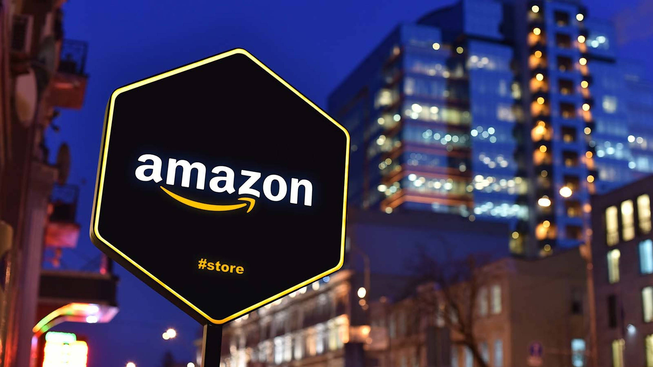 Amazon 街道標誌 壁紙 — 夜間，一個發光的六邊形Amazon商店標誌矗立在模糊的城市建築前。