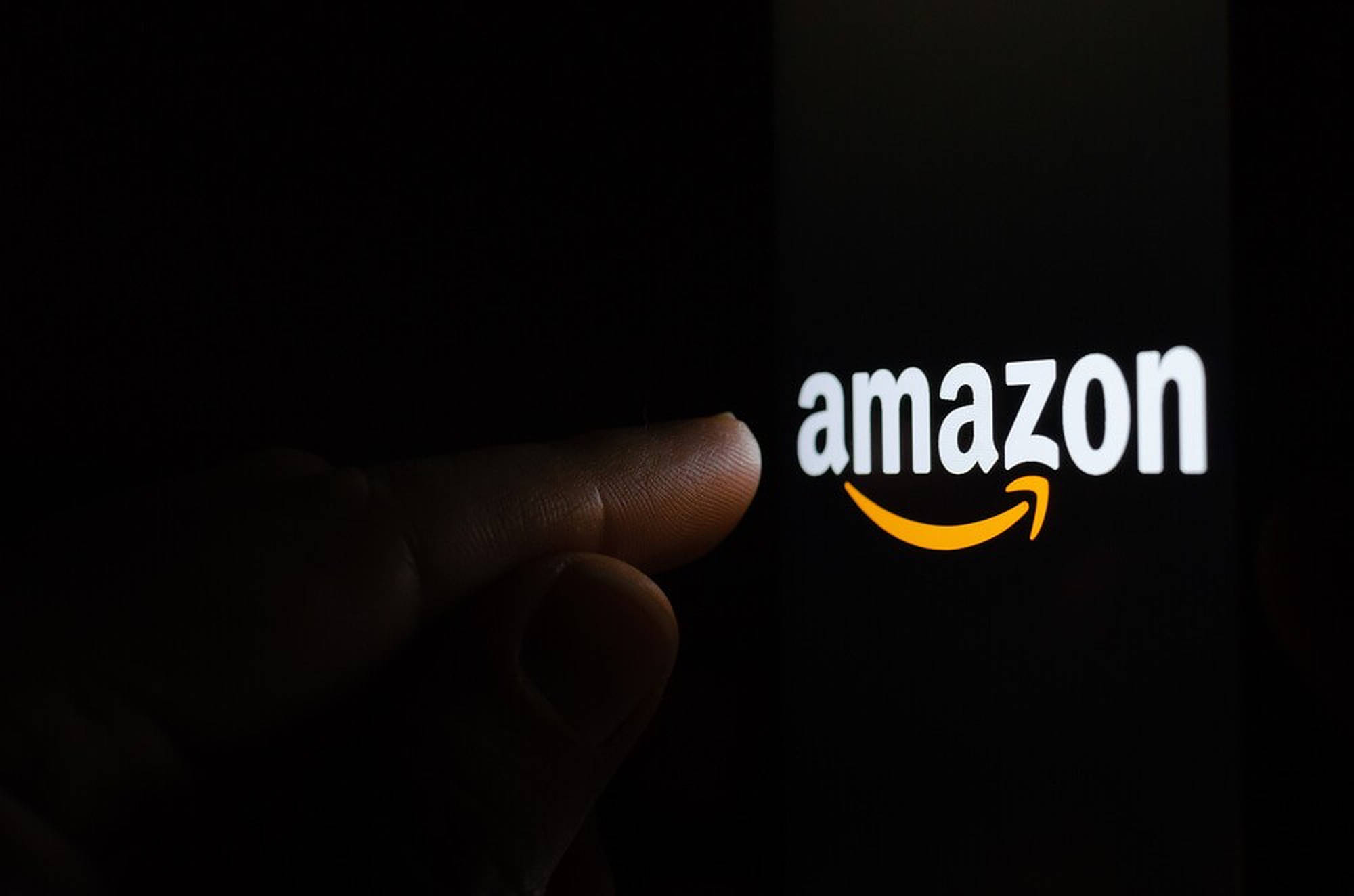 Amazon 觸控螢幕 壁紙 — 在昏暗的環境中，一隻手指點擊黑暗智能手機屏幕上發光的Amazon標誌。