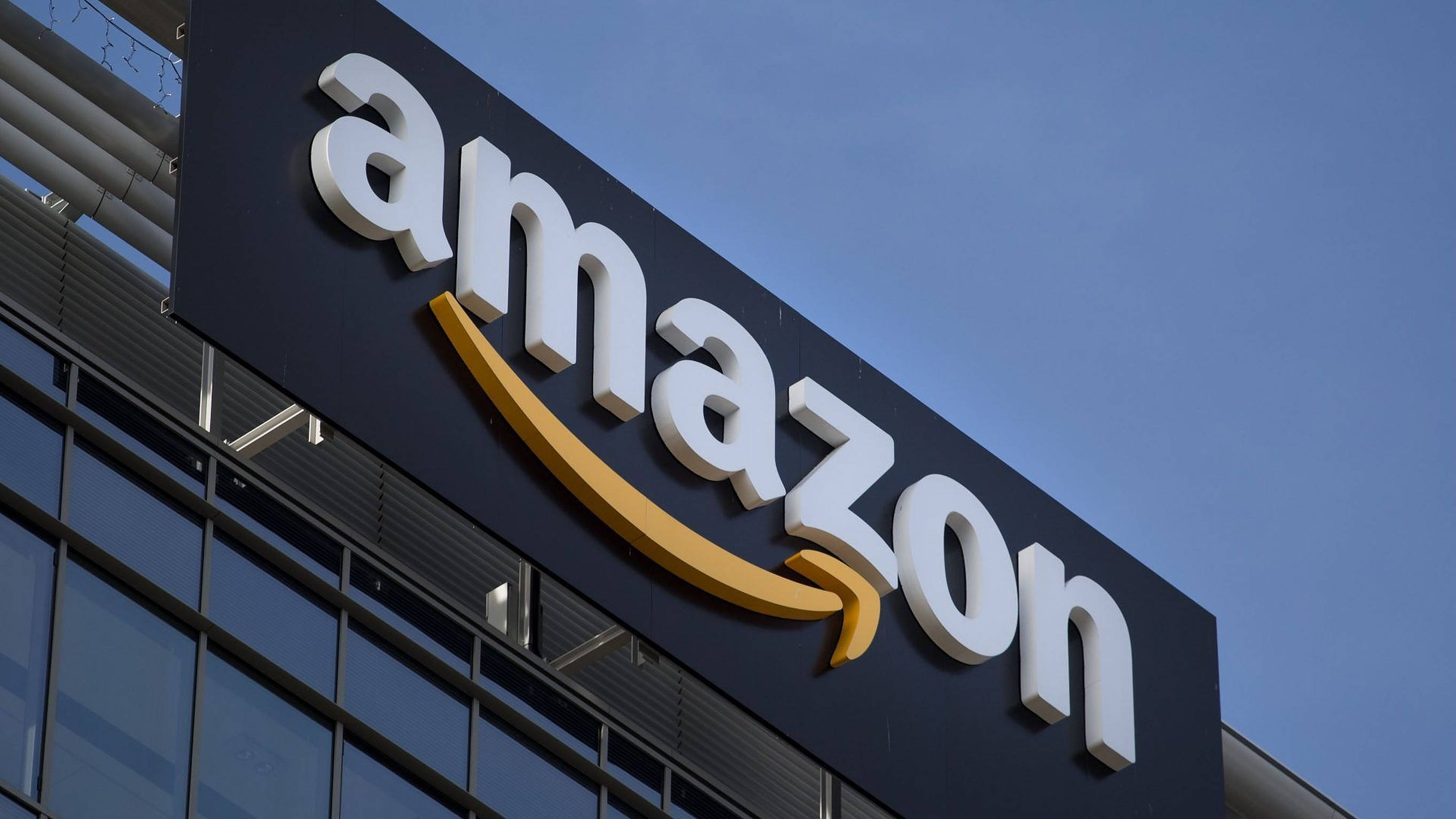 Amazon標誌於大樓 壁紙 — 特寫亞馬遜標誌，白色和金色，安裝在現代玻璃建築上的深色標牌上，背景是晴朗的藍天。