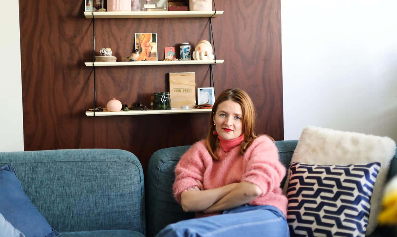 Amélie Pichard在沙發上享受悠閒時光 壁紙 — 一位穿著粉紅色毛衣的女性自信地坐在藍色梳化上，梳化上有圖案靠墊，背景是木質層架牆。
