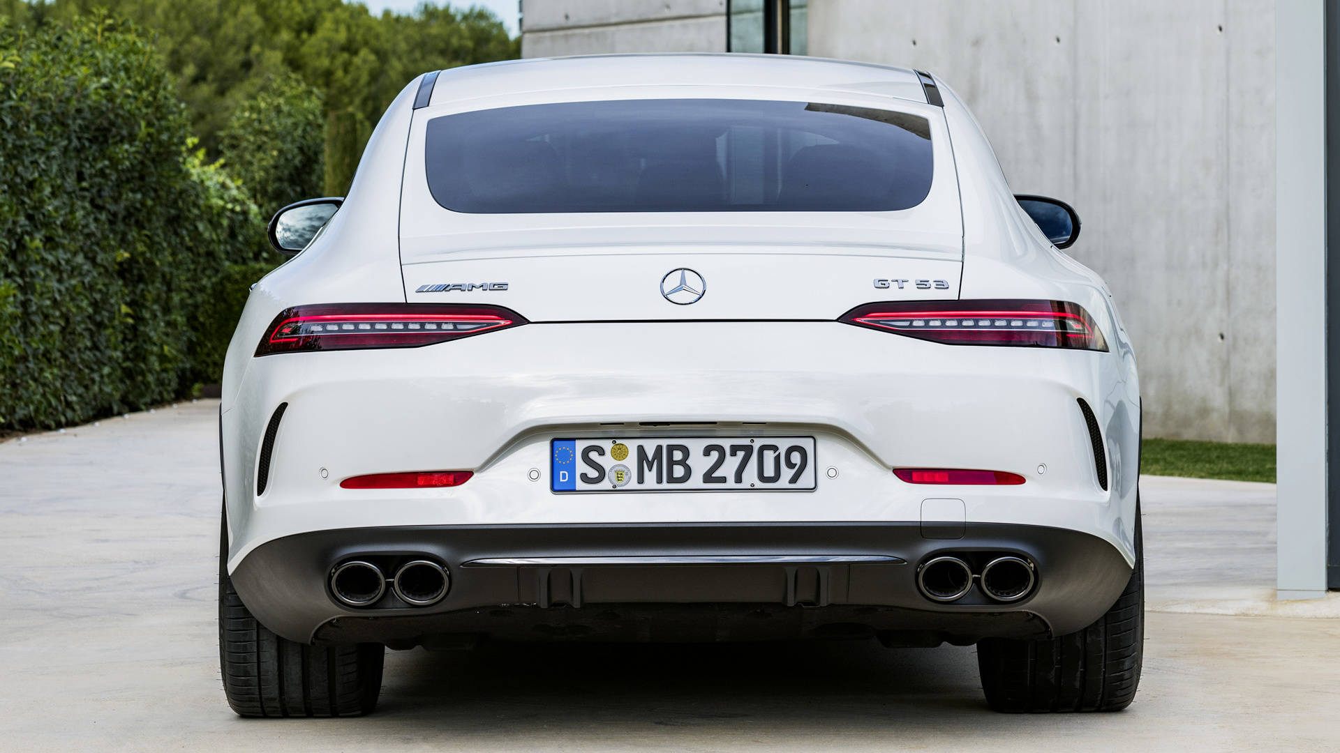 梅賽德斯-AMG GT 53 壁紙 — 白色Mercedes-AMG GT S的後視圖，停放在混凝土車道上，旁邊有綠色樹籬和現代建築。