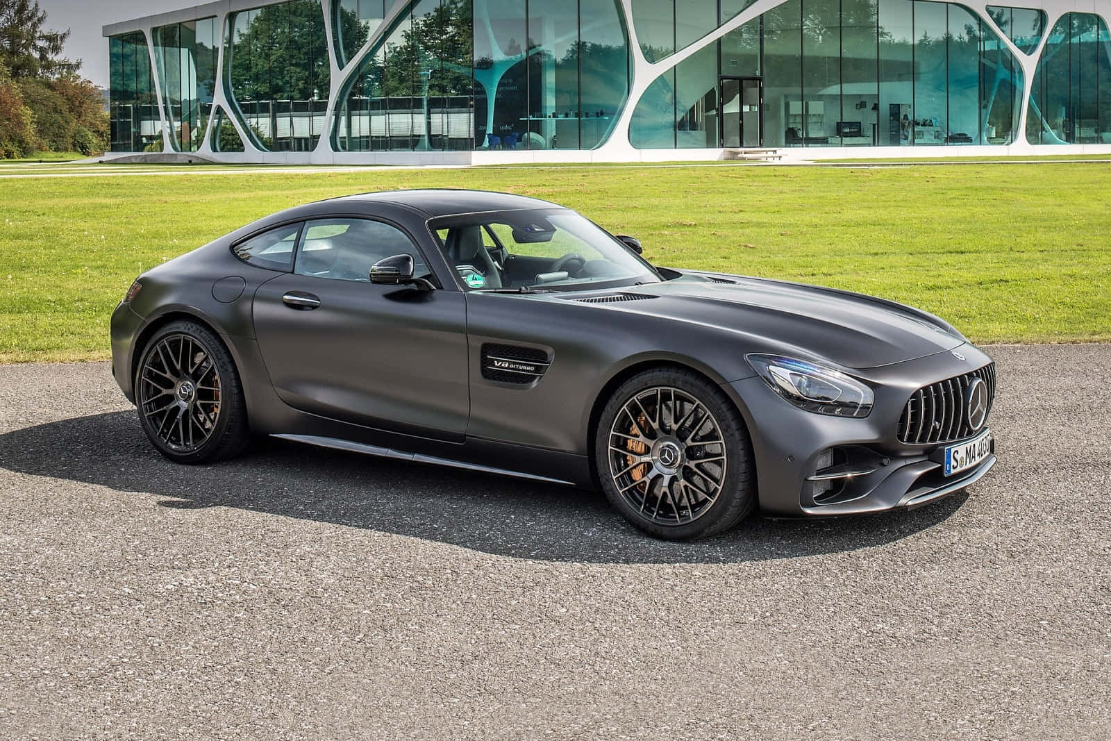 準備以時尚姿態駕馭道路：最佳 AMG GT-R 壁紙 — 一架啞光灰色的 Mercedes-AMG GT 跑車停泊在瀝青路上，前面係一棟有綠色草地嘅現代玻璃建築。