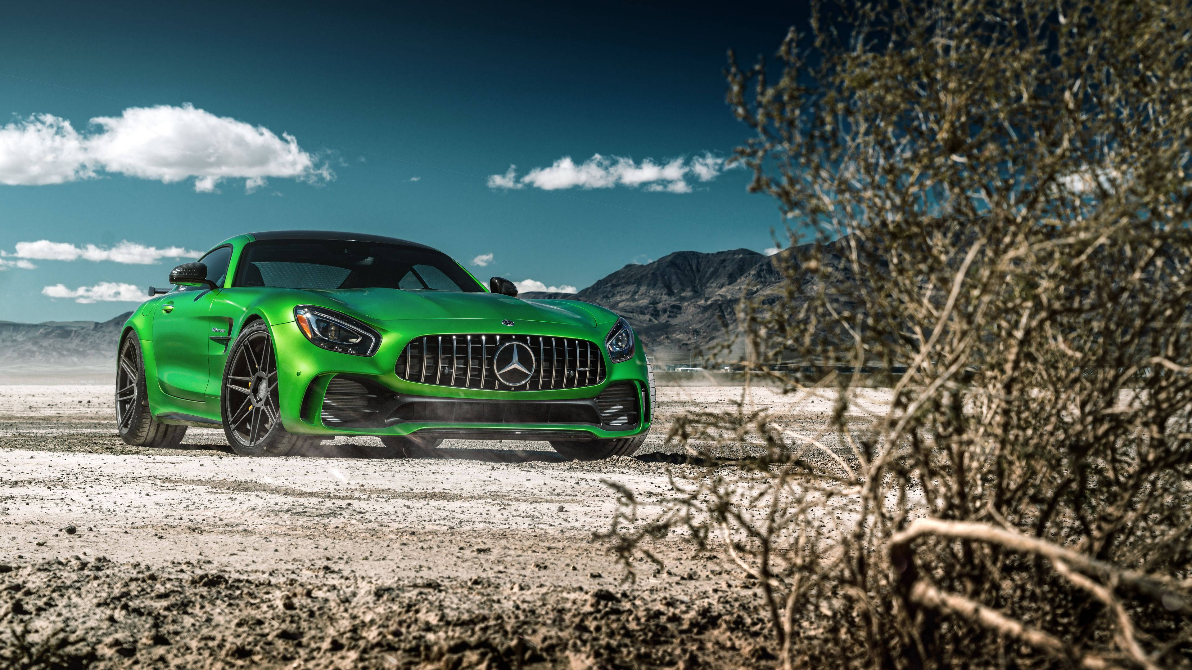 AMG GT R 深綠天空 壁紙 — 一輛充滿活力的綠色Mercedes-AMG GT跑車停泊在塵土飛揚的沙漠道路上，背景有山脈和乾燥灌木叢，藍天白雲。