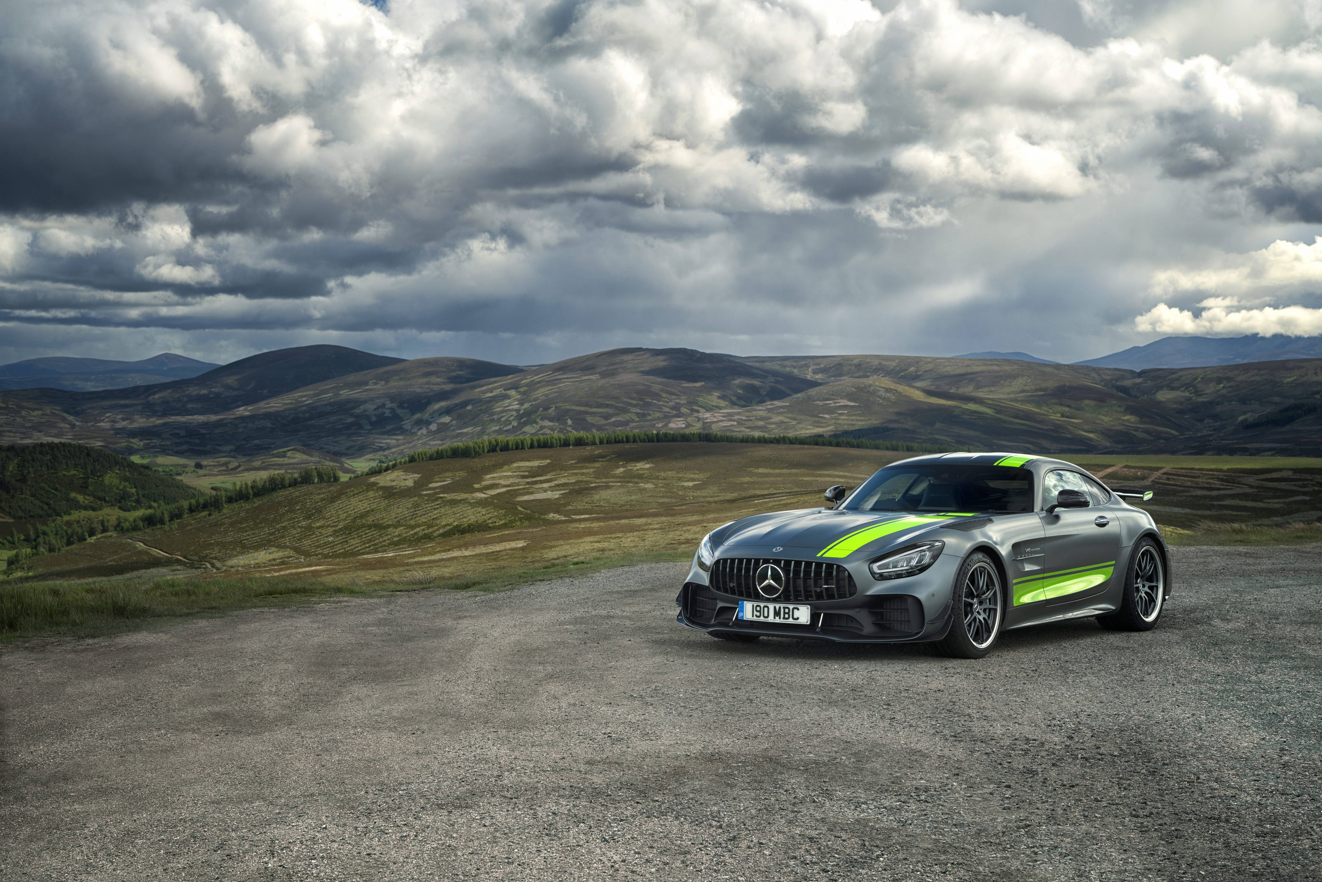 銀綠 AMG GT R 壁紙 — 一輛時尚的銀色 Mercedes-AMG GT 帶有螢光綠色條紋，停泊在碎石路上，背景是戲劇性的多雲天空和起伏的丘陵。