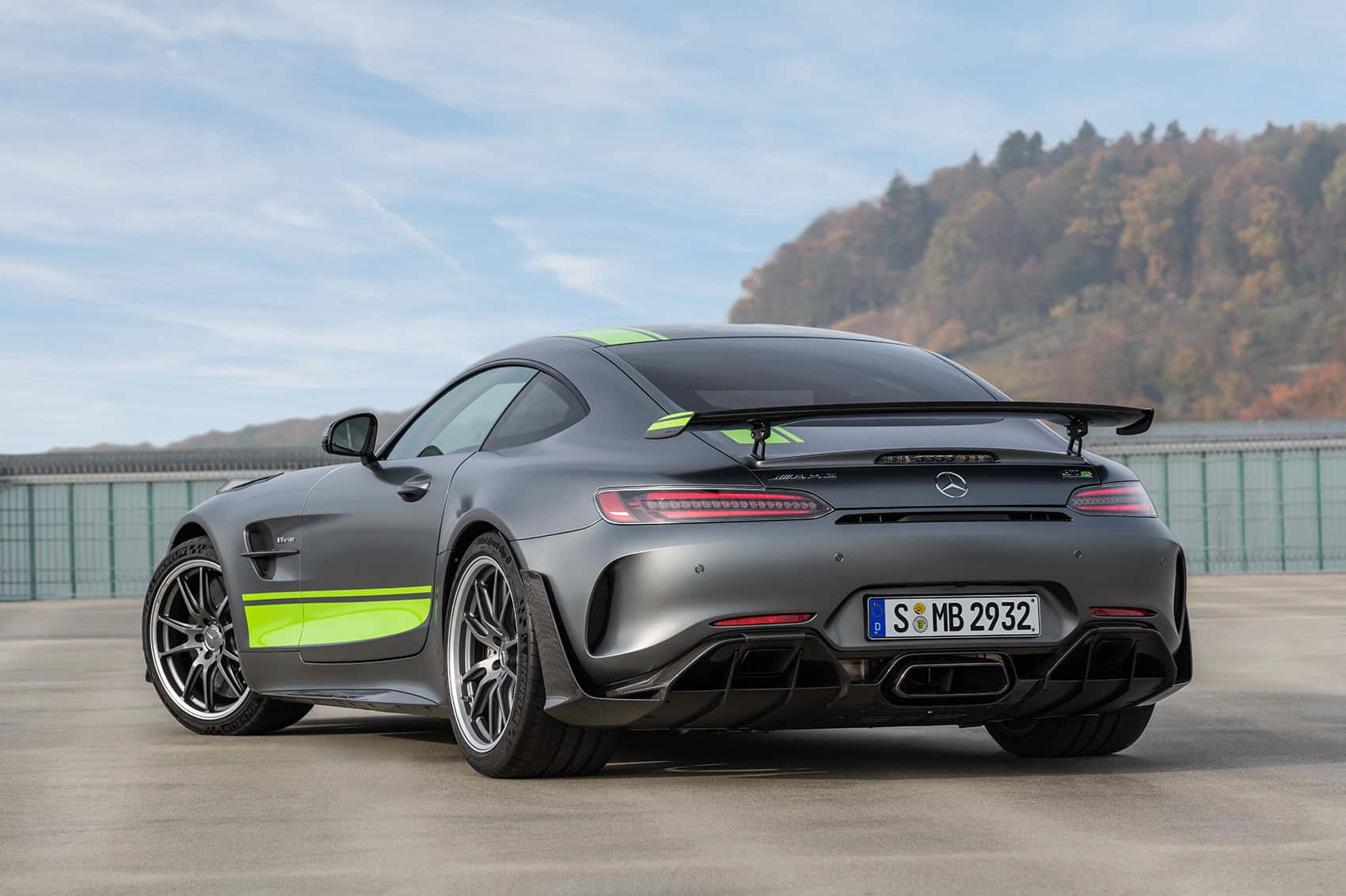 最佳 AMG GT-R Pro 綠色條紋背景 壁紙 — 一輛啞光灰色的 Mercedes-AMG GT R 跑車，帶有螢光綠色點綴，停放在混凝土表面上，背景是森林覆蓋的山丘，天空部分多雲。