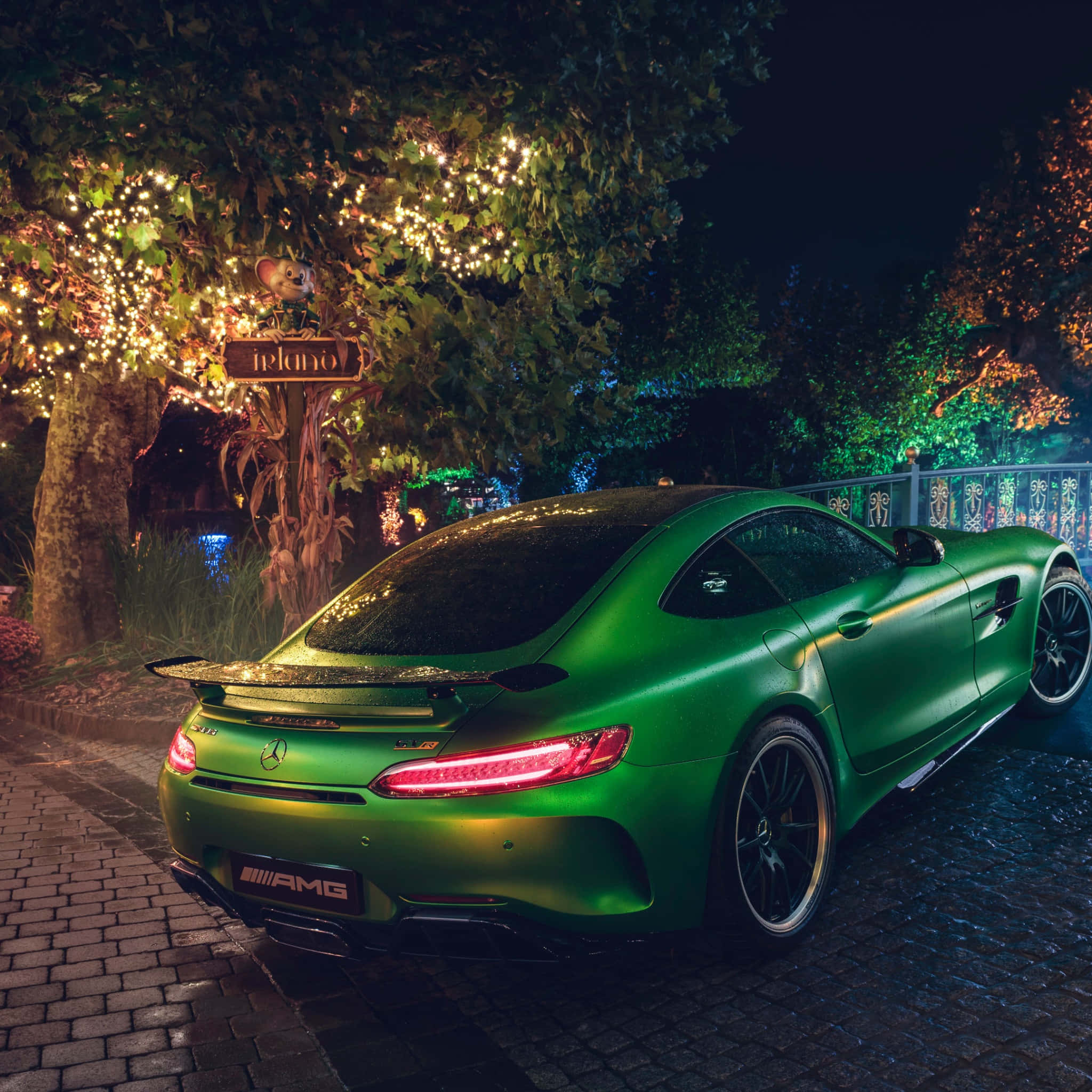最佳 AMG GT-R 夜間後視背景 壁紙 — 一架啞光綠色的 Mercedes-AMG GT 在夜晚停泊在發光的串燈下，位於鵝卵石小徑上。