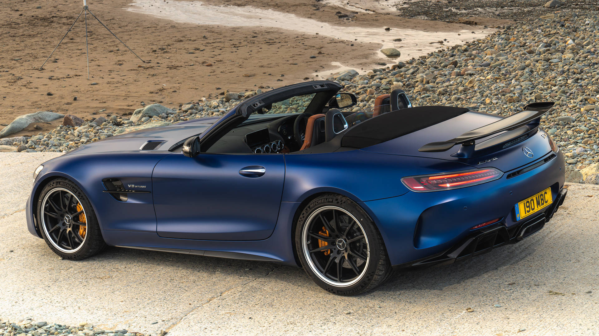 深藍色AMG GT R 壁紙 — 一架啞光藍色Mercedes-AMG GT Roadster停泊在岩石海灘旁邊的混凝土板上，背景有一個三腳架。