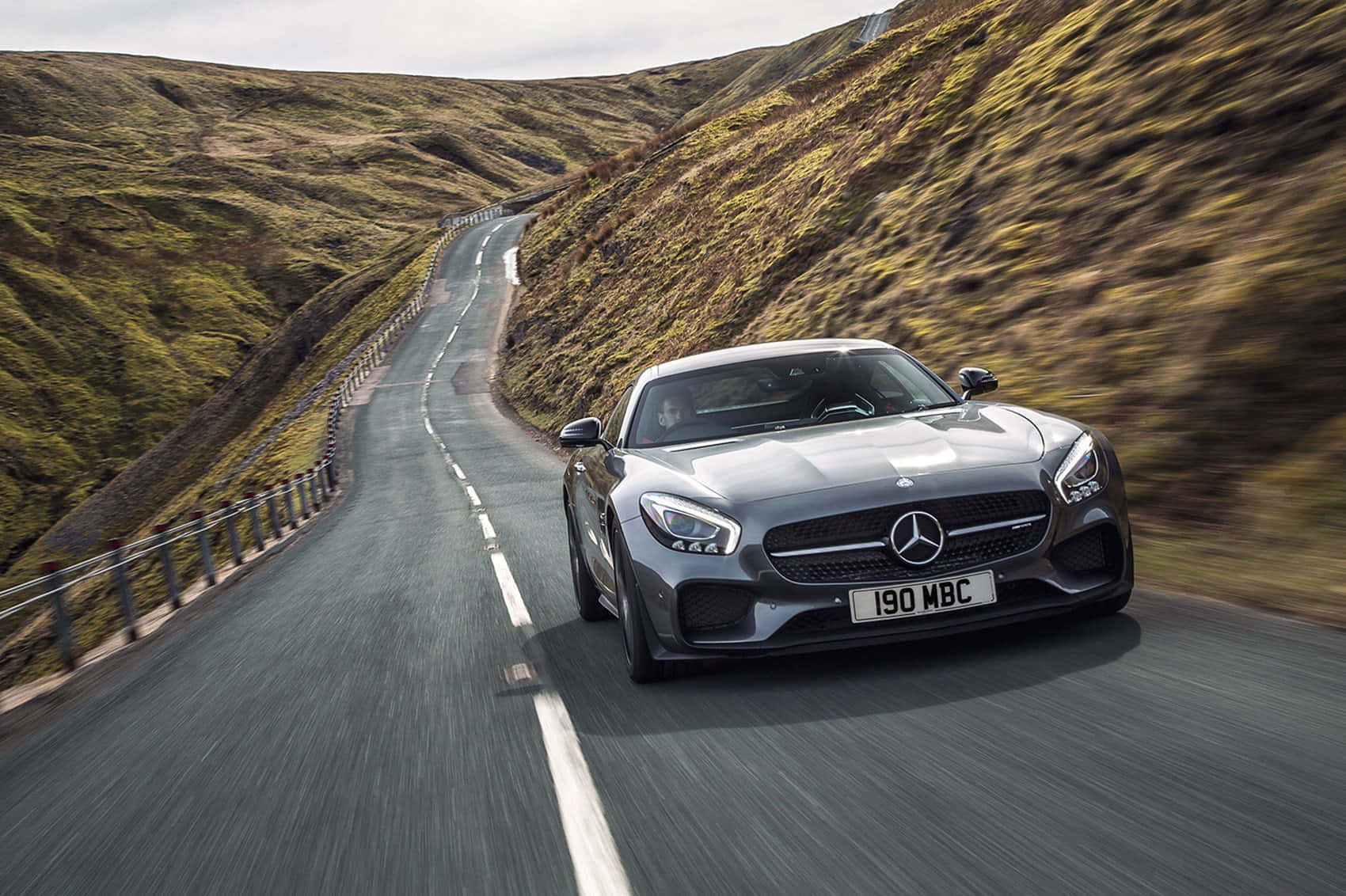 令人屏息的靈活駕馭：震撼AMG GT-R背景 壁紙 — 一架流線型銀色 Mercedes-AMG GT 在陰天之下，沿著蜿蜒的山路穿過翠綠的山丘飛馳。