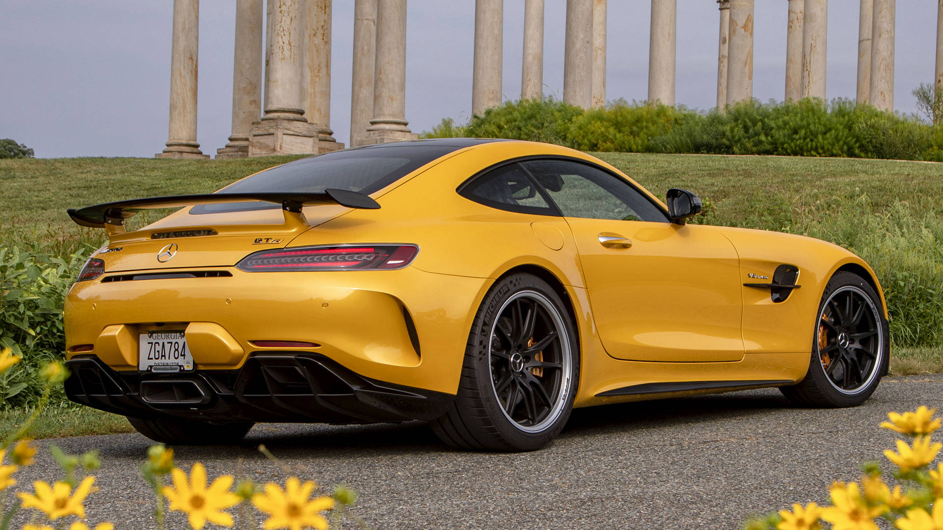 黃色 Mercedes-AMG GT R 停泊在古老石柱附近，前景有黃色花朵。